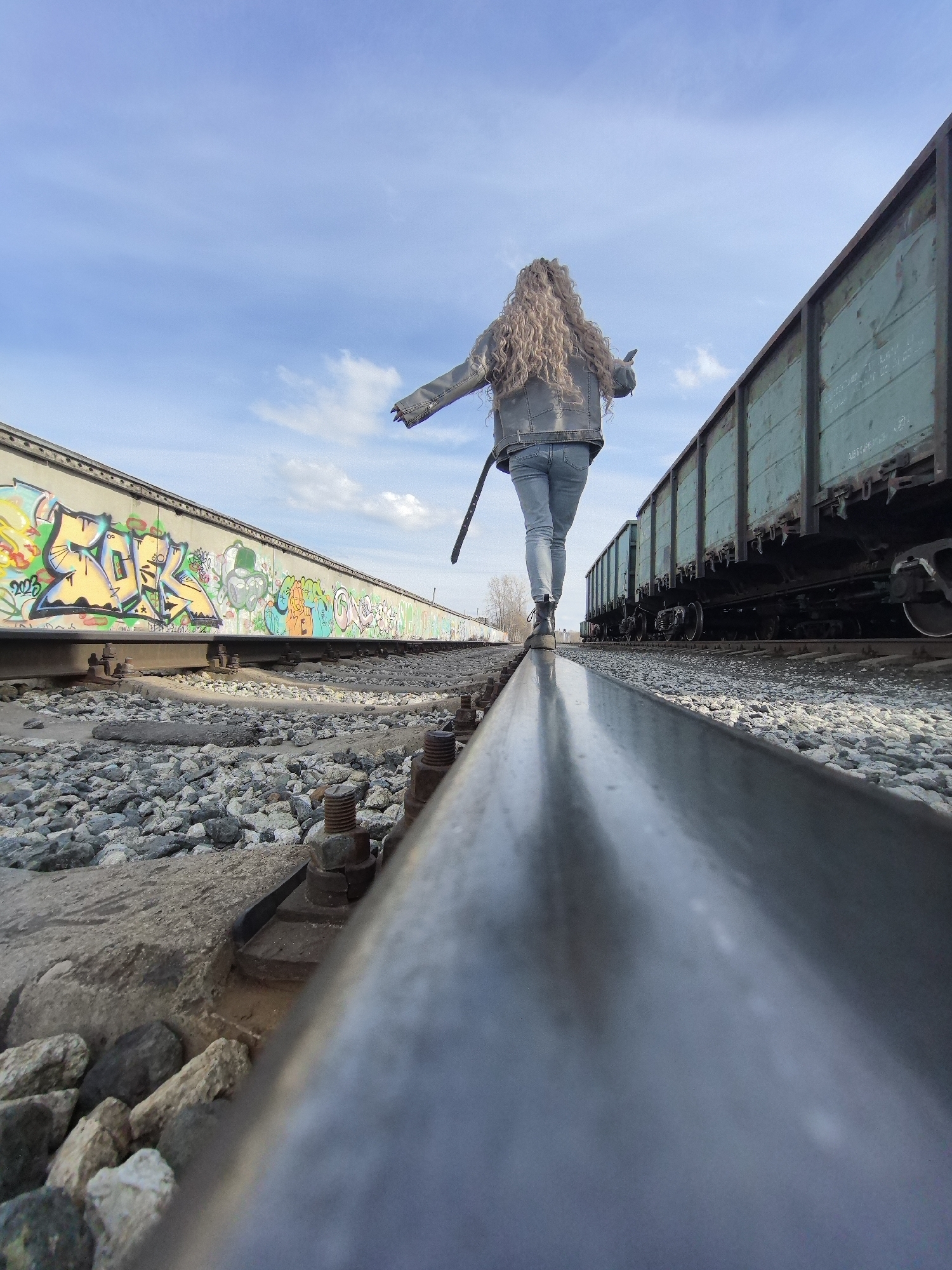 The image depicts a person walking on a railroad track. The location is in the city of Chaikovsky, Russia. The person, seen from the back, has long curly hair and is wearing a denim jacket, jeans, and black boots. They are balancing on the rail, with arms extended slightly to the sides. On either side of the track are parked railroad cars. One car has graffiti on it, and the other is a faded green color. The ground around the tracks is covered in gravel. The sky is blue with some clouds. The photo is taken at a low angle, emphasizing the rail in the foreground.