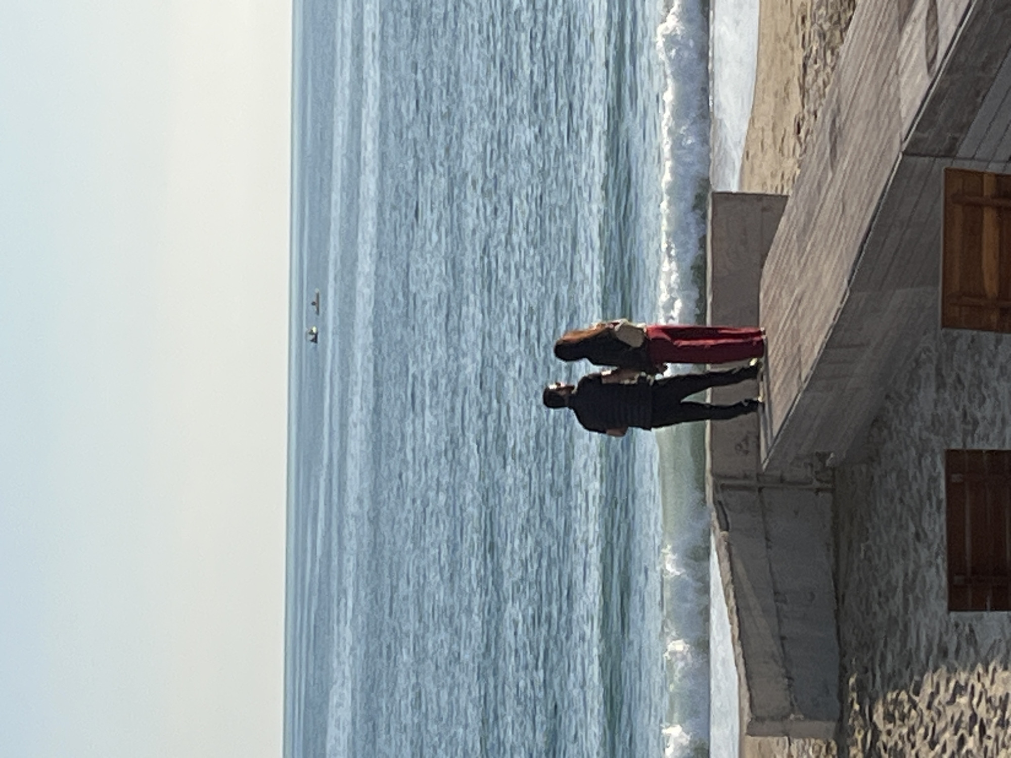A medium shot depicts a man and a woman standing on an elevated stone pier or wall overlooking the sea in Alanya, Turkey. The man, closer to the camera, wears a black shirt and pants. The woman, slightly behind him, wears a black jacket and red pants. They both appear to be looking out at the sea. The pier or wall is made of grey stone and features wooden window shutters on its right side. The sea is a textured blue, with white waves crashing near the pier. In the distance, two birds are visible in the sky. The sky itself is a pale blue, almost white.