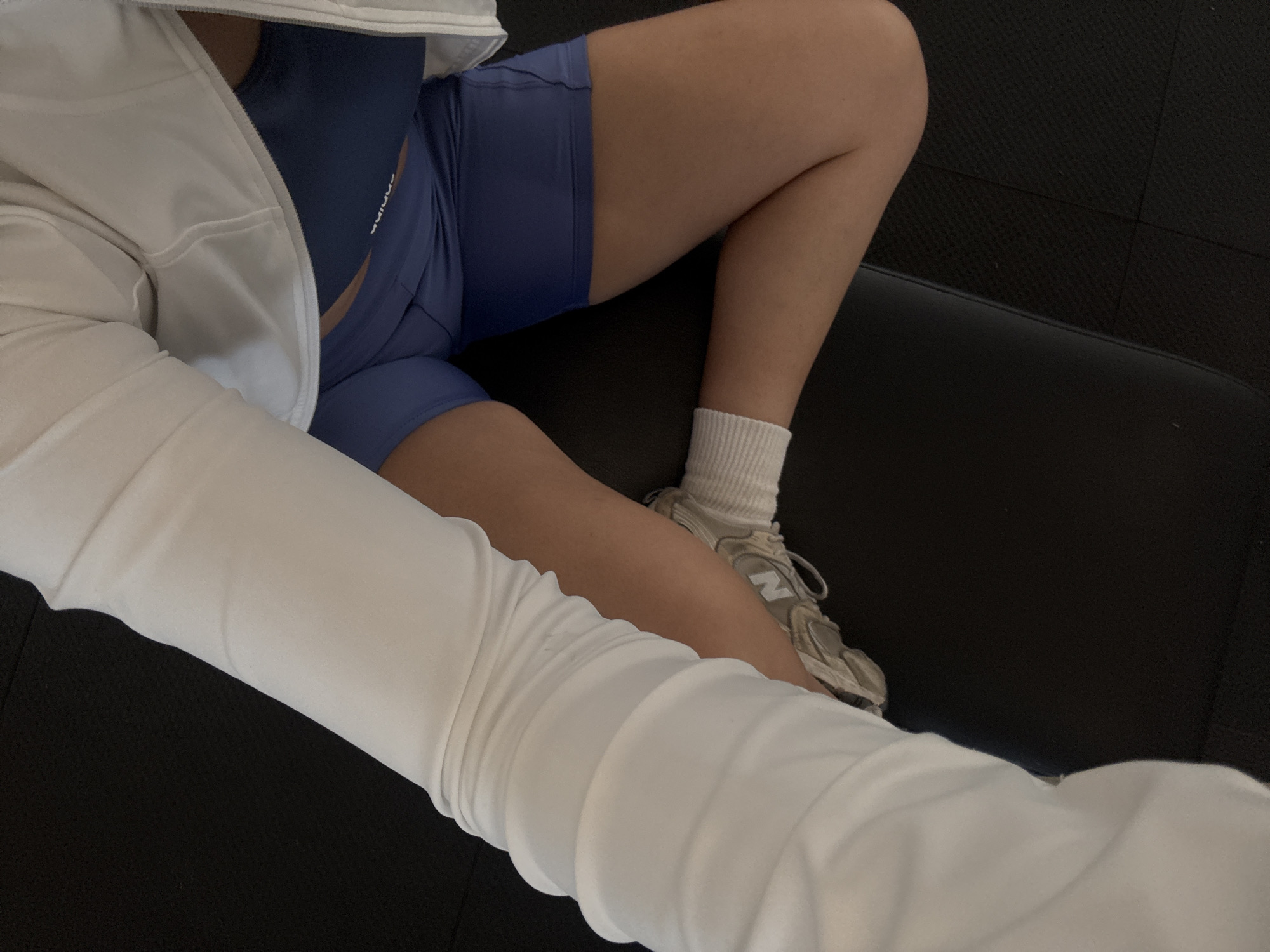 A close-up, high-angle shot shows the lower body of a person sitting on what appears to be a dark, possibly black, upholstered surface. The person is wearing a blue athletic bra and matching shorts. A white zip-up jacket covers the upper body and part of the arm. One leg is visible, with a white sock and gray New Balance sneaker on the foot. The background is dark, likely a part of the seating arrangement. The location cannot be determined from this image alone.