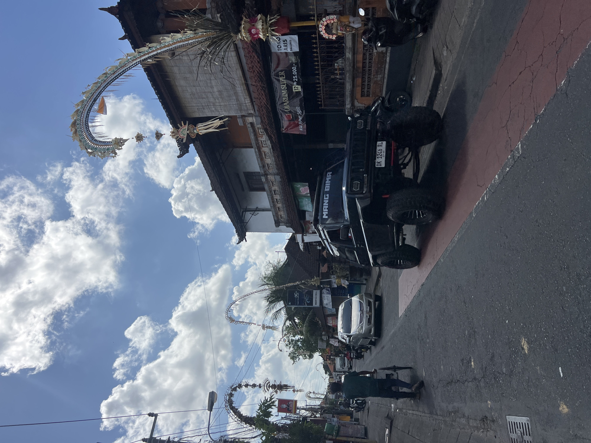 An outdoor, street-level, eye-level, full shot captures a busy road in North Kuta, Indonesia. A black, lifted, off-road vehicle dominates the foreground. The vehicle's license plate reads "DK 1246 RL." To the left of the vehicle, a person walks away from the camera, pushing a cart. Further down the street, a silver sedan is visible. The street is lined with buildings, some adorned with traditional Balinese decorations, including elaborate arches strung across the road and floral arrangements. Power lines and street lights are visible. The sky is blue with scattered white clouds. The scene suggests a bustling urban environment with cultural elements.