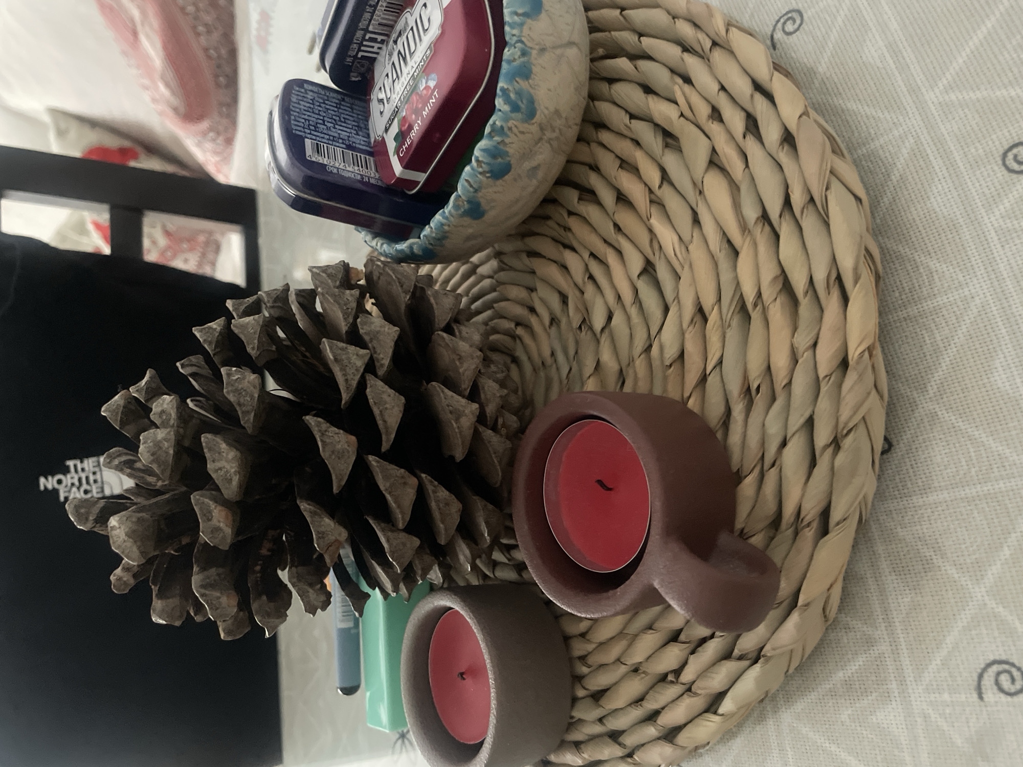 The image depicts a still life arrangement on a table in a domestic setting, possibly in Atarfe, Spain. The primary focus is a large pinecone positioned centrally on a woven placemat. To the left of the pinecone, two small, red tealight candles in rustic holders are visible. One holder is a simple, cylindrical brown ceramic, and the other is a dark red ceramic with a handle. To the right of the pinecone, a small, decorative bowl with a blue rim contains several tin containers, including one labeled "SCANDIC CHERRY MINT." In the background, out of focus, are a black item that appears to be a bag or jacket with "THE NORTH FACE" branding, and a piece of furniture with cushions that have a red and white patterned fabric. A green marker pen is partially visible behind the pinecone and to the left. The overall impression is of a curated display of natural and decorative items.