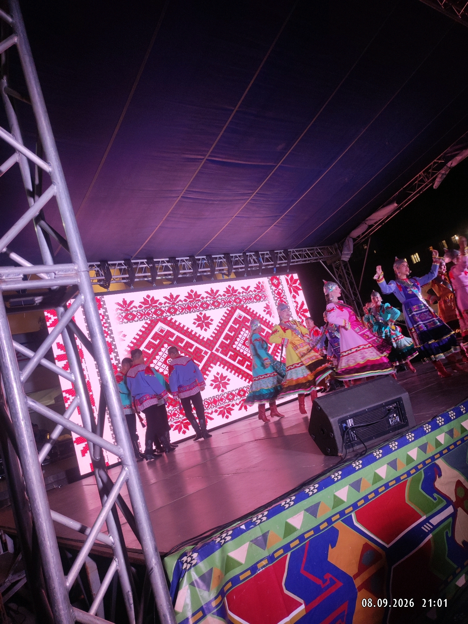 A stage performance is underway at an outdoor event. Several individuals are on stage, some dressed in traditional colorful attire. In the background, a large LED screen displays a red and white embroidered pattern. To the left, a metal scaffolding structure is visible. A large speaker sits on the stage. The overall environment suggests a festival or cultural event. The lower right corner of the image displays the date "08.09.2026" and time "21:01".