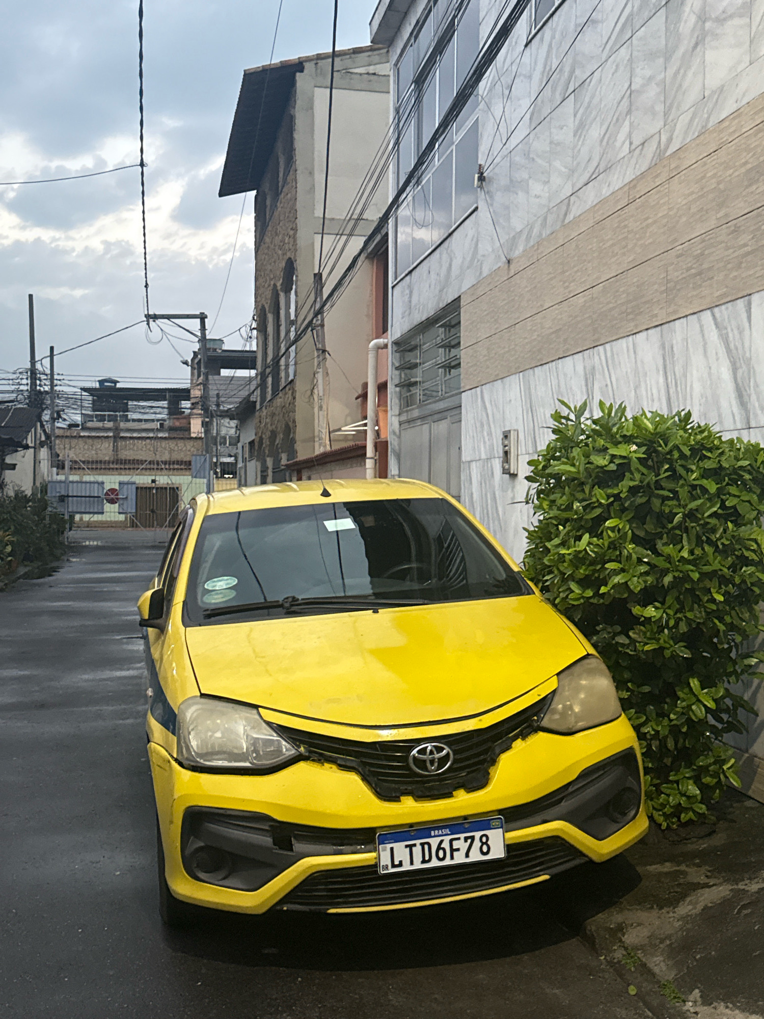 A yellow taxi with the license plate LTD6F78 is parked on a wet asphalt street in Nilópolis, Brazil. The car is a Toyota Etios and it is facing forward, slightly angled to the right. The street is narrow and bordered by buildings on both sides. To the right of the car is a large green bush. The buildings are of various architectural styles and appear to be older. The sky is overcast, suggesting it may have recently rained or is about to. The scene is an urban street with a mix of residential and commercial structures.