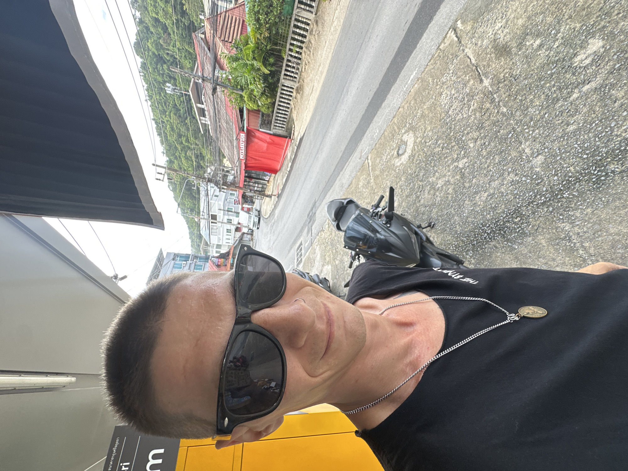 A man is standing on a sidewalk in Karon, Thailand, taking a selfie. He is wearing sunglasses, a black t-shirt, and a silver chain with a pendant. Behind him, a black motorcycle is parked. The background shows a street with buildings and lush green hills. The scene appears to be in a tropical or sub-tropical region due to the dense foliage on the hills. The sky is overcast.