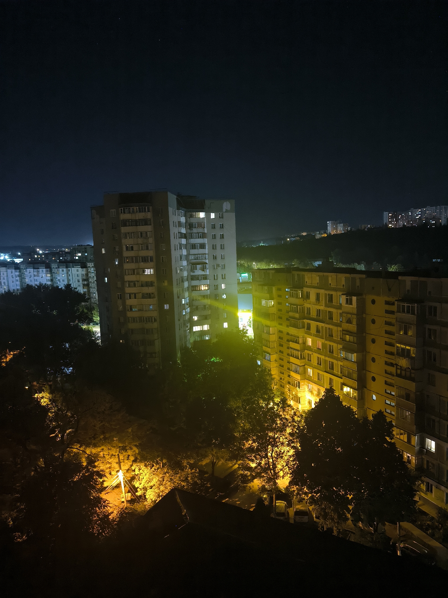 The image captures a nighttime view of a residential area in the city of Chișinău, Moldova. Dominating the scene are several multi-story apartment buildings, their windows illuminated with a warm, yellow light, suggesting occupancy. The architecture appears to be typical of Soviet-era construction, with long, rectangular blocks of apartments. Trees, silhouetted against the artificial lights, line the streets and surround the buildings, creating a dense, dark foreground in some areas. A street light is visible, casting a bright yellow glow on the surrounding vegetation. In the distance, more buildings and lights can be seen, indicating the urban sprawl of the city. The sky is dark, with a few faint stars visible, suggesting a clear night. No people or specific actions are discernible. The overall impression is one of a quiet, residential neighborhood at night, with the warm glow of apartment lights contrasting with the dark expanse of the night sky and the shadows of the trees. The specific neighborhood is likely in a peripheral district of Chișinău, characterized by these types of high-rise apartment blocks and surrounding green spaces.