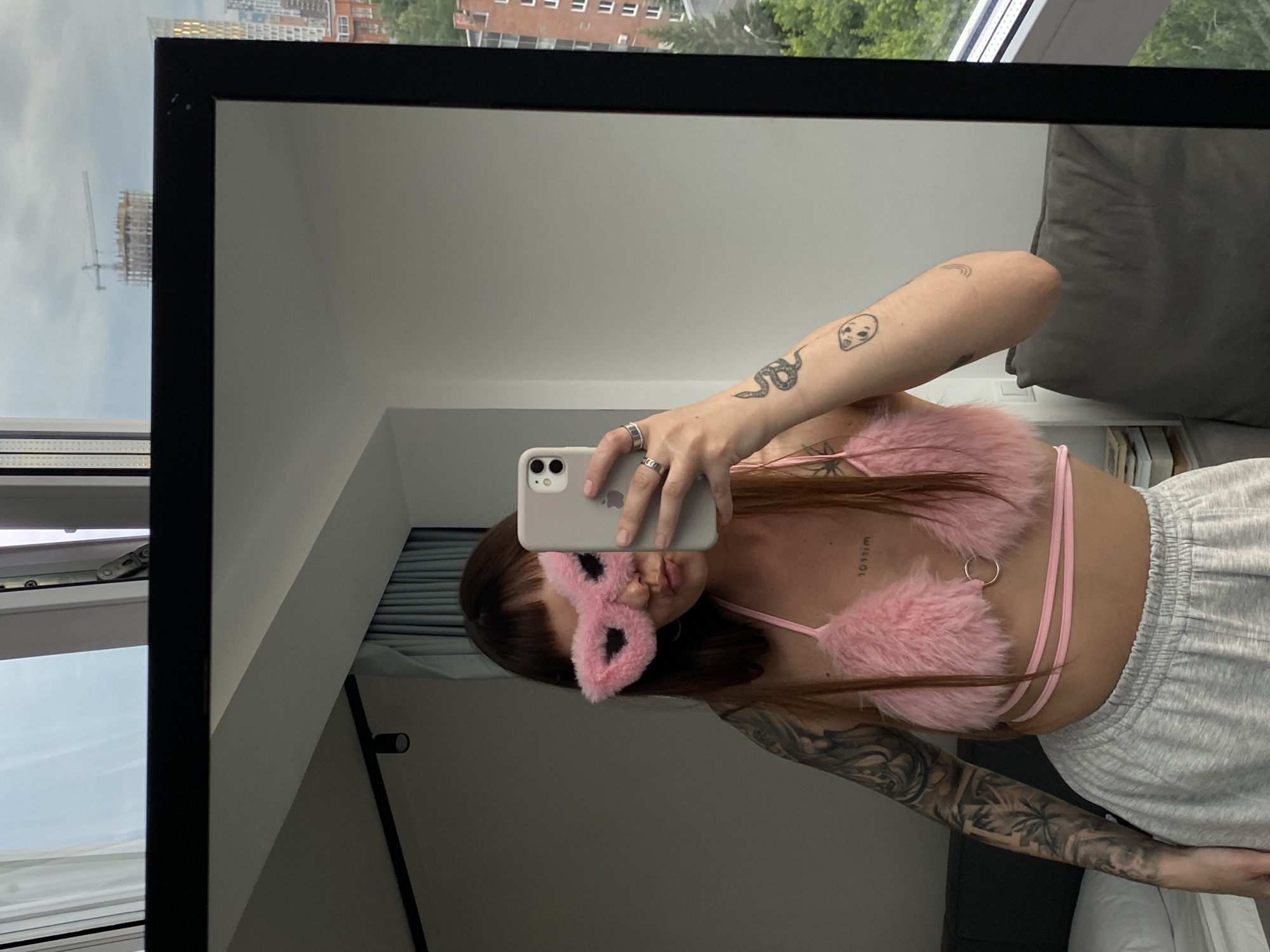 The image, taken in Izhevsk, Russia, shows a woman holding a phone in her left hand, taking a selfie in a mirror. She is wearing a pink fluffy bra and grey sweatpants, and a pink fluffy mask over her eyes. Tattoos are visible on her arms and chest. The background shows a window with a view of buildings and trees, as well as a partially visible bed and pillow. The overall mood appears casual and relaxed. The time of day and weather are not explicitly discernible. There is some text visible on her chest, but it is difficult to read.