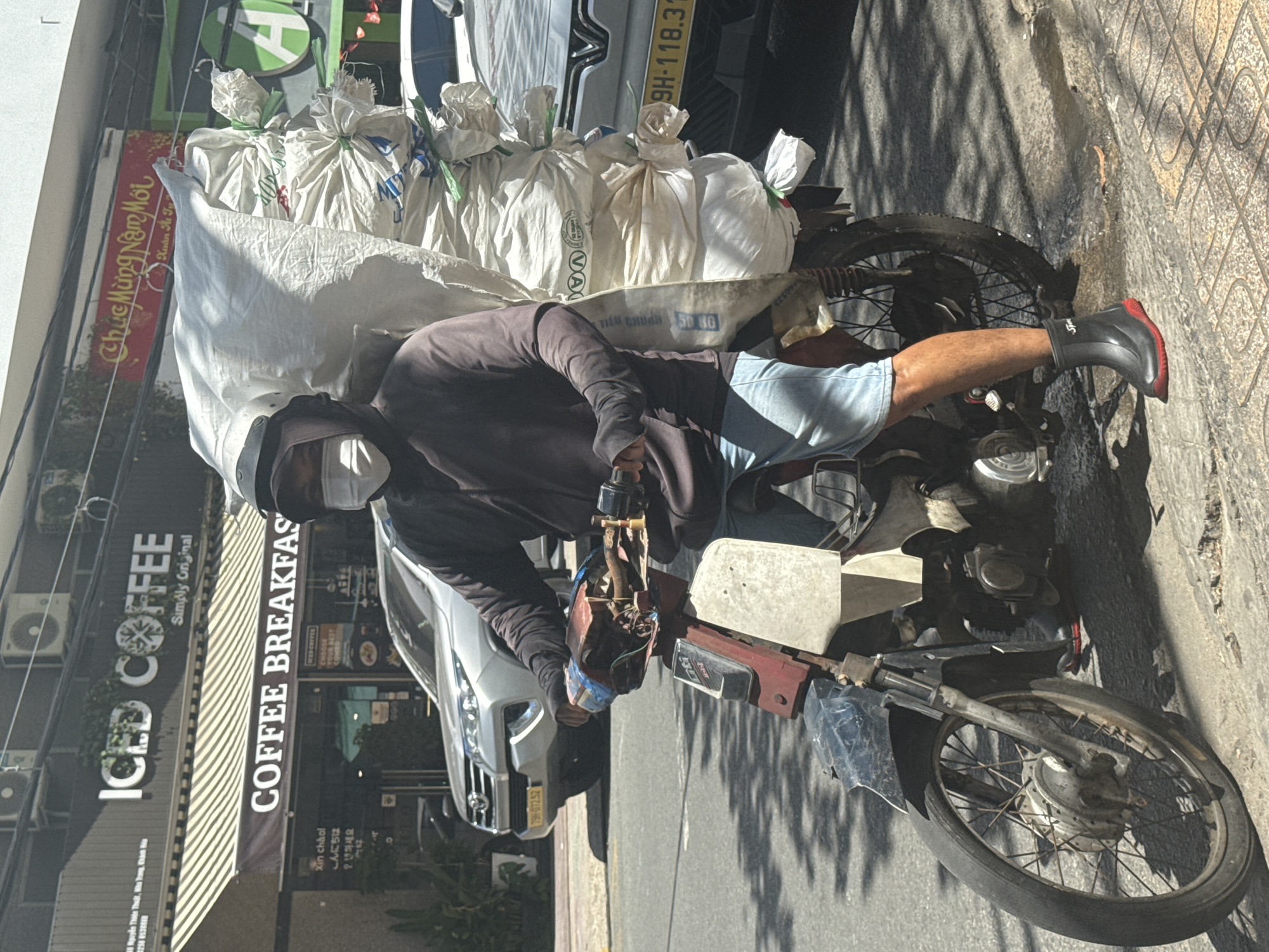 Here's a description of the image:
The photograph shows a man riding a motorbike in what appears to be a city street. The motorbike is heavily loaded with several large white sacks. The man is wearing a dark hooded jacket, light blue shorts, a face mask, and black boots with red soles. He's focused on operating the motorbike.
In the background is a building with signs indicating a coffee shop. One sign reads "ICED COFFEE" and "COFFEE BREAKFAST". There are also Vietnamese characters on the sign above, and other foreign languages below. Cars are parked on the street, and one of the cars has the license plate "9H-118.31". The light suggests it's a sunny day, as evidenced by the shadows on the street.