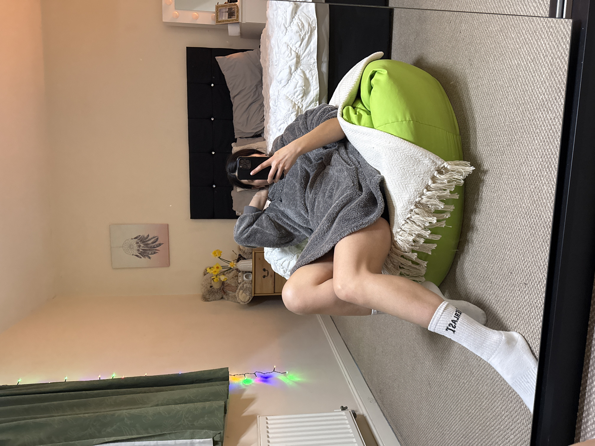 The image depicts an interior bedroom scene captured via a mirror selfie. A female individual, partially obscured by a black smartphone, is seated in a bright green bean bag chair, positioned on a beige textured carpet. She is wearing a grey, fuzzy, long-sleeved garment and white socks with black, reversed text "T2AJAS" visible on the right sock. Her right hand holds the smartphone, angled to capture the reflection, while her left hand rests behind her head.
The room features light peach-colored walls. A large bed with a black upholstered headboard and white quilted bedding is central to the background. To the left of the bed, a two-drawer wooden bedside table holds a large brown teddy bear and a white vase containing yellow artificial flowers. To the right of the bed, a white vanity mirror with illuminated bulbs is present, next to a small golden-framed picture. Along the left wall, a white radiator is installed below a dark green patterned curtain. A string of multi-colored LED fairy lights hangs vertically on this wall. No visual cues within the image confirm the specific location as Great Yarmouth, United Kingdom.
