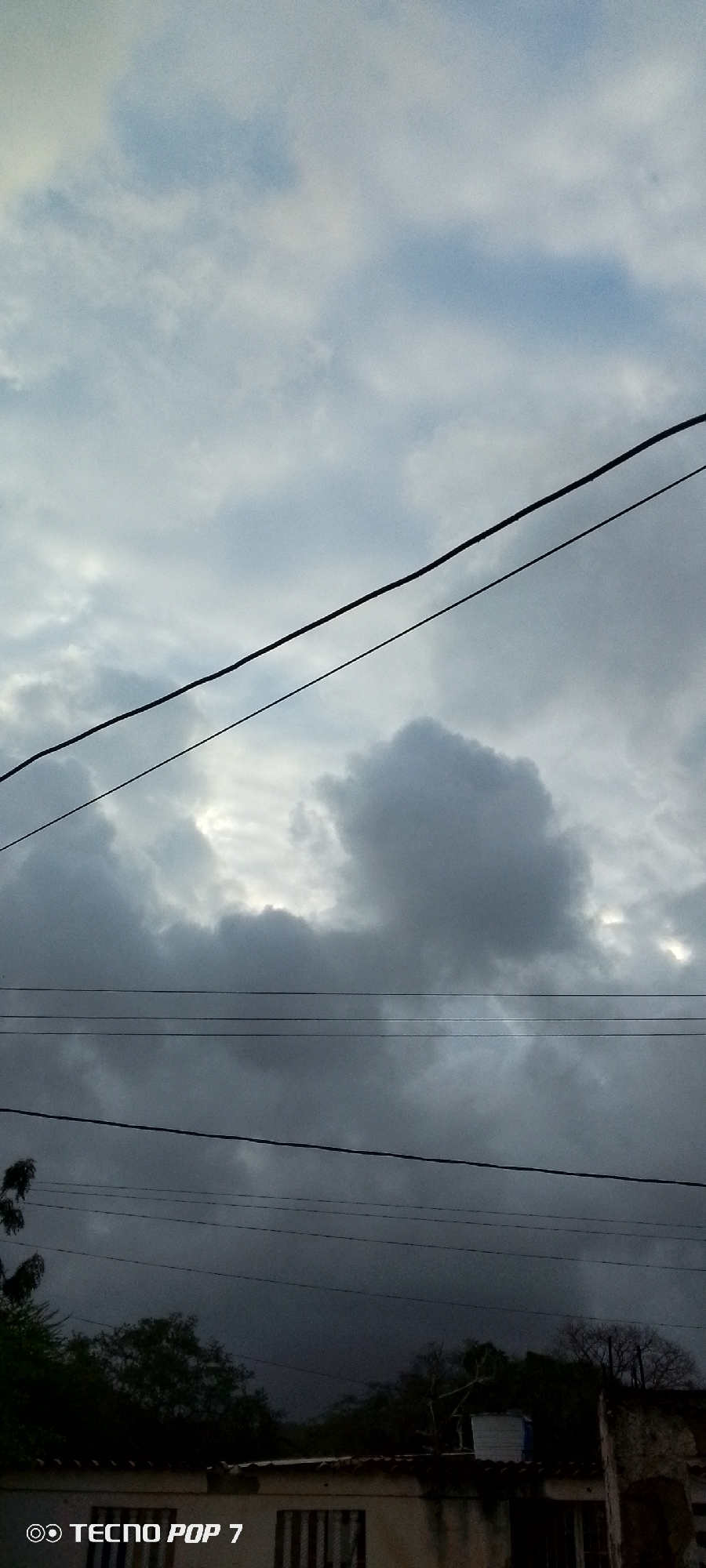 This is a vertical, close-up shot of a cloudy sky with power lines in Altagracia, Venezuela. Three dark, horizontal power lines are visible across the frame, spaced at varying intervals. The sky is filled with a mix of light and dark clouds, with patches of brighter sky peeking through. In the lower portion of the image, the tops of trees and a building roof are visible, silhouetted against the dark clouds. The building appears to be made of concrete with striped window coverings. The "TECNO POP 7" watermark is in the bottom left corner.