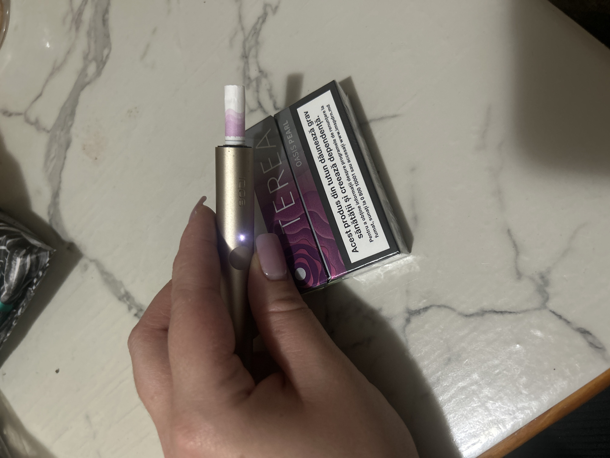 An overhead shot displays a hand with a lilac-painted fingernail holding a gold and silver e-cigarette device. The device is inserted into a pack of "TEREA" cigarettes, which are branded with a purple floral pattern and feature text in Romanian that translates to "This product is harmful to your health and causes addiction." The pack is placed on a marble-patterned surface, which shows some subtle stains and smudges. In the background, a blurred, dark object is partially visible. The scene is illuminated by artificial light, casting soft shadows. The specific location in Basarabeasca, Moldova, is not determinable from the visual evidence provided.