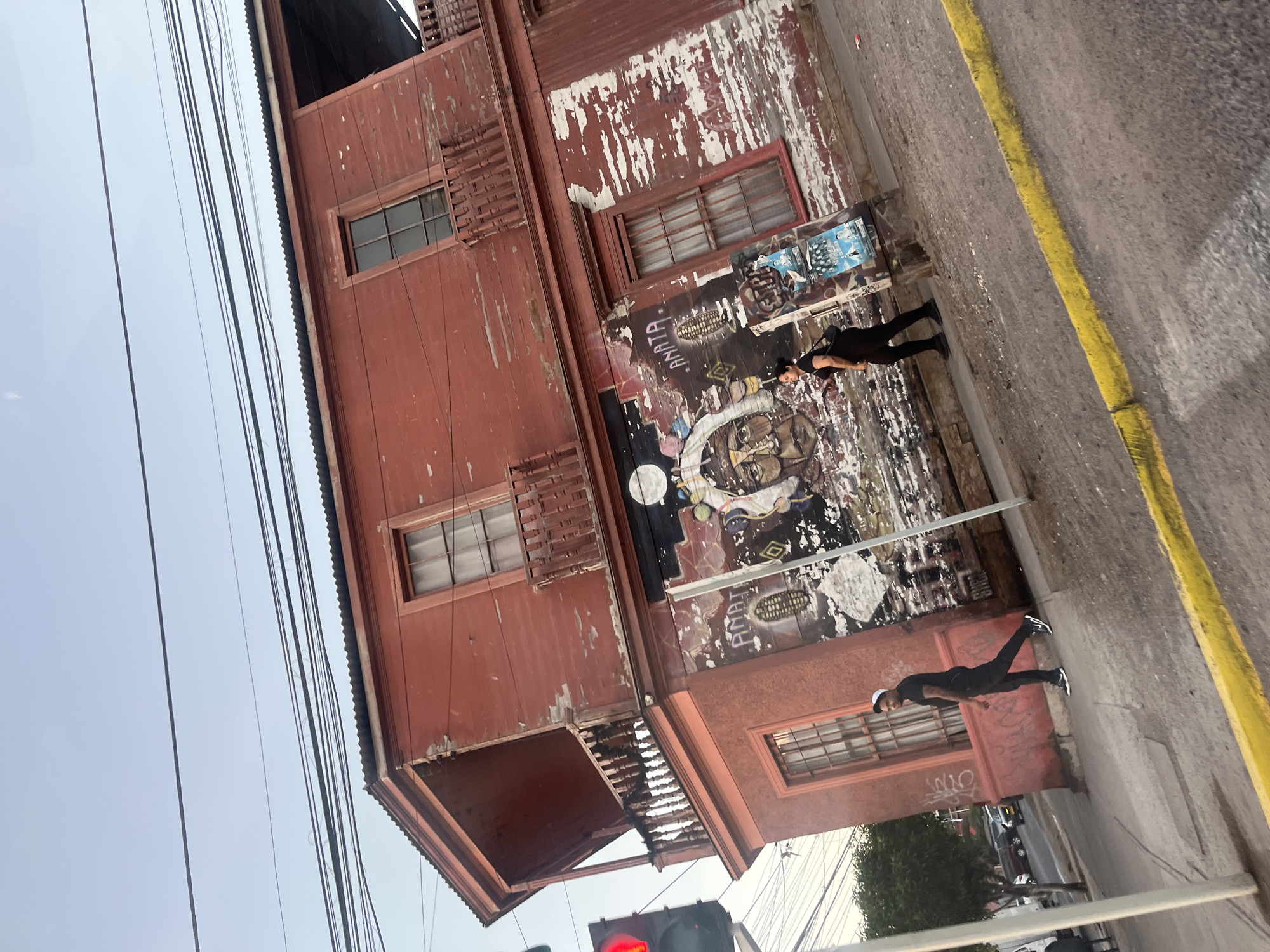 The image depicts a street in an unidentified city, possibly in South America, based on the architectural style. A three-story red building dominates the left side of the frame. The facade is visibly aged, with peeling paint and signs of wear and tear. The building features multiple windows with dark frames and ornamental window shades. The building has an overhang and balcony.
The building's exterior wall is covered with graffiti, posters, and various street art. Two individuals are standing on the sidewalk in front of the building. One is closer to the camera and is seen wearing a black outfit and sneakers. The second person is further back, also dressed in black, and wearing a hat.
A yellow line marking a curb runs along the right side of the image, adjacent to the sidewalk. The sky is visible in the upper left corner, with power lines running parallel to the building. In the lower part of the image, a traffic light can be seen with its red light on. Cars are parked on the street in the lower right of the frame.