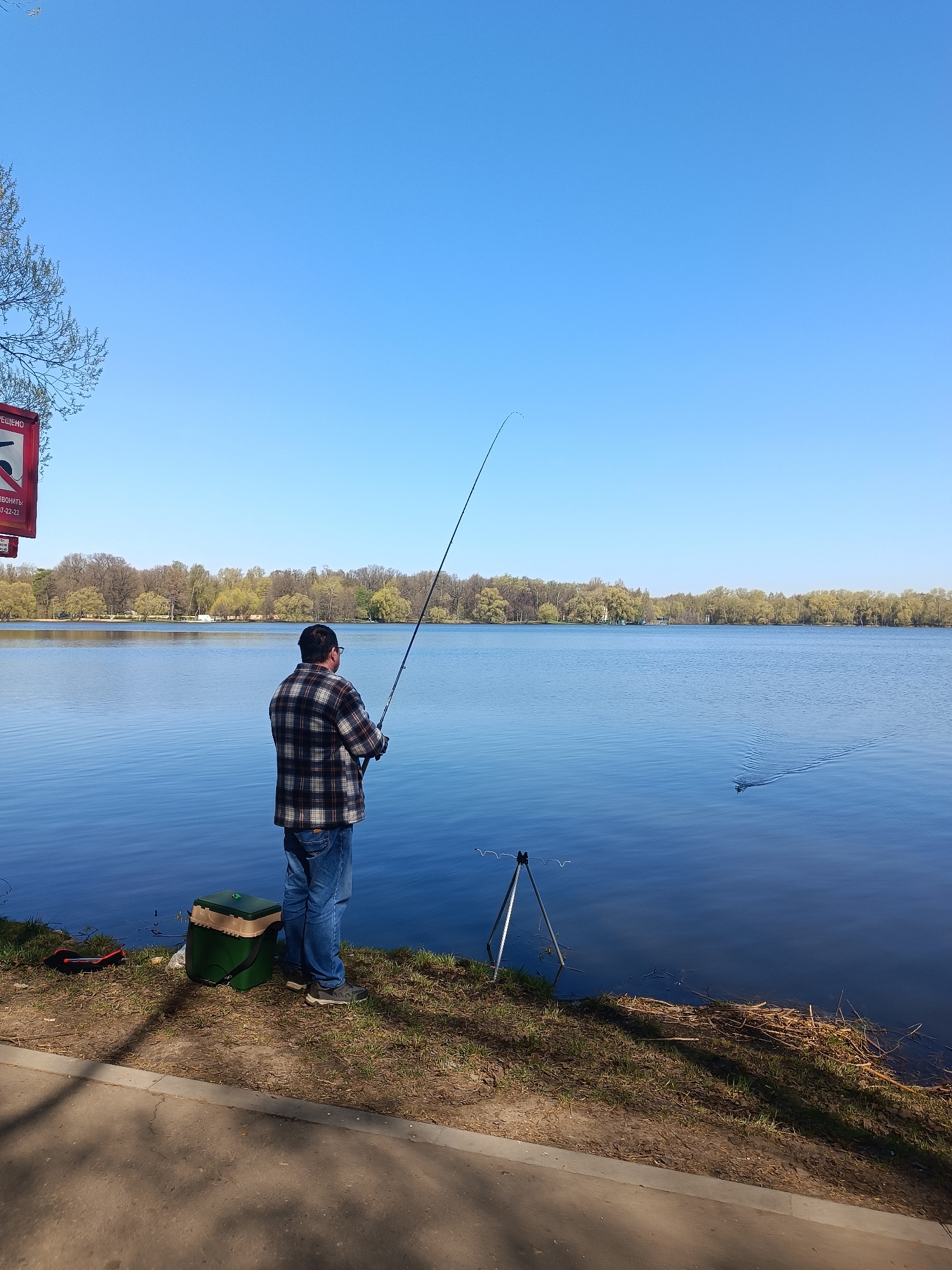 A man stands fishing on the bank of a body of water on a sunny day. The man, wearing a plaid shirt, jeans, and a cap, holds a fishing rod. Beside him is a green fishing tackle box and a rod stand. The water is calm with a slight ripple, and the opposite bank is lined with trees. A sign is visible on the left, with text in Russian and some red coloring. The location is likely in Moscow, Russia, as indicated by the Russian language on the sign. A concrete sidewalk runs along the edge of the bank where the man is standing.