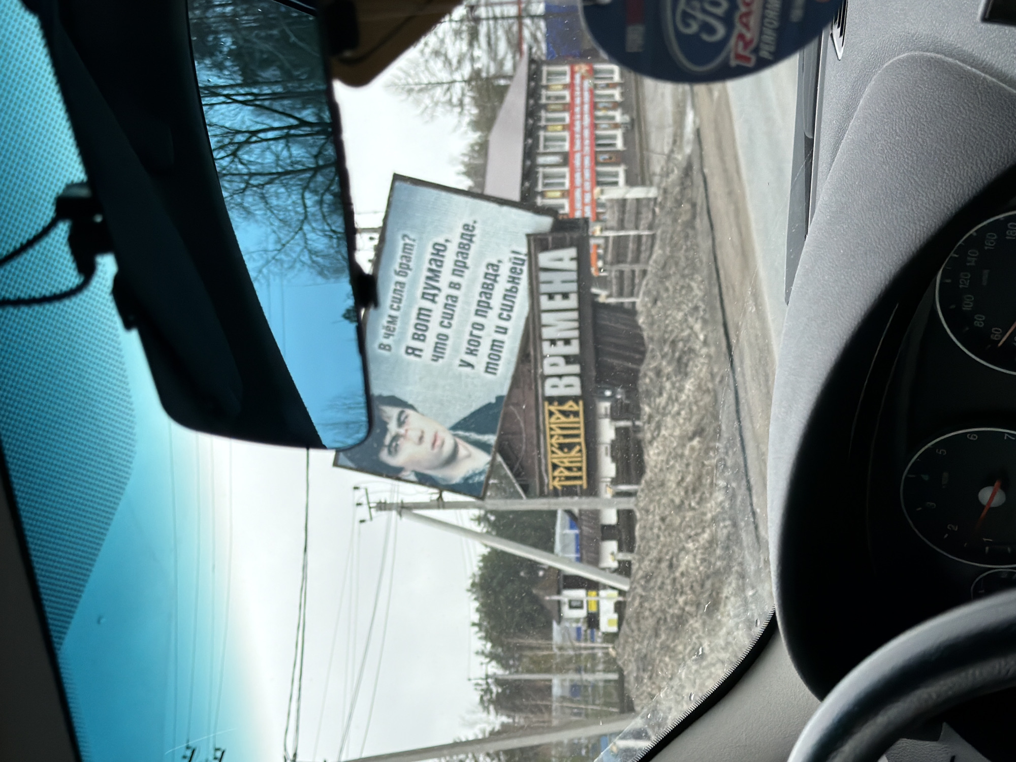 The image shows a view from inside a car, likely a Ford, with the steering wheel and dashboard partially visible. The camera is focused on a billboard advertising a company called "ТРАКТИКЕ ВРЕМЕНА." The billboard features a man with a dark haircut and an injured face, and includes the following Russian text: "В чём сила брат? Я вот думаю, что сила в правде. У кого правда, тот и сильней!" The scene appears to be on a street in Noviy, Russia, with buildings and some vegetation visible in the background. The weather seems overcast.