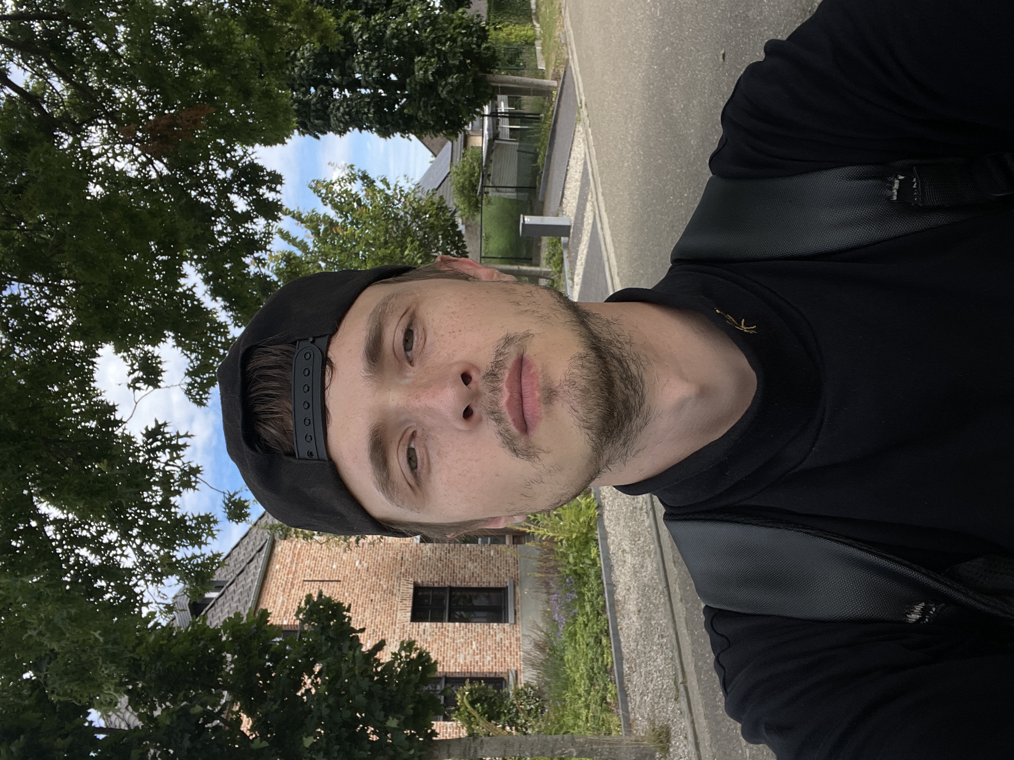 A medium shot selfie is taken outdoors in Diepenbeek, Belgium, on a paved road with a tree-lined background. The subject is a young Caucasian male with light skin, brown eyes, and short brown hair, wearing a black baseball cap and a black turtleneck shirt. He has a light beard and mustache and a few freckles on his face. He is looking directly at the camera with a neutral expression. He is also wearing a black backpack. In the background, there is a red brick house with black-framed windows, a garden with green plants and purple flowers, and several trees. Further in the distance, there are more houses with solar panels on their roofs and a trash bin on the side of the road. The sky is blue with some white clouds.