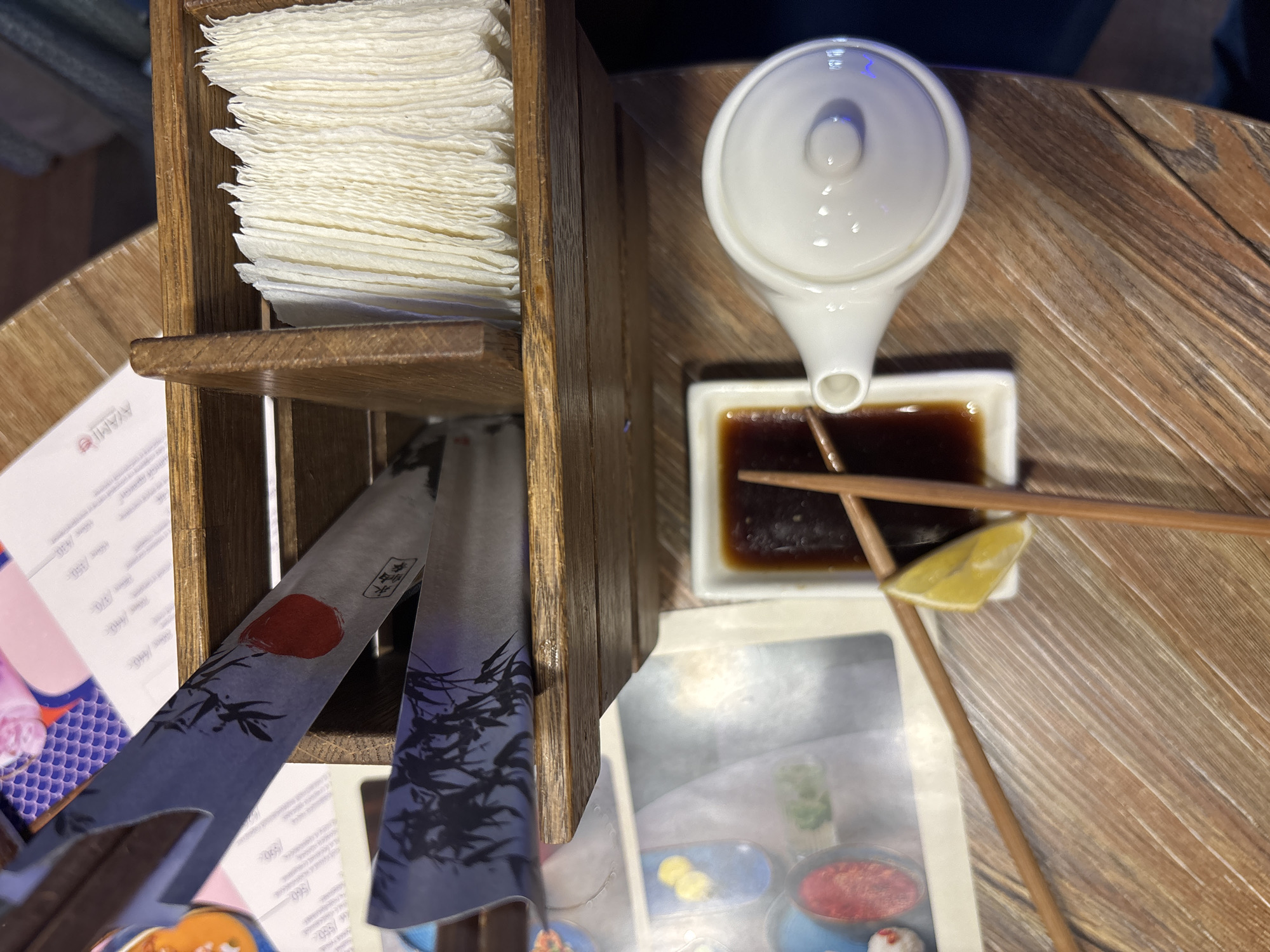 The scene captures a close-up, overhead view of a dining table in a restaurant, identified as likely AMI Izhevsk in Izhevsk, Russia, based on visible menu details. The table is made of light brown wooden planks. A multi-compartment wooden caddy is centrally located, containing a stack of white paper napkins in one section and individually wrapped chopsticks in another. The chopstick wrappers are blue/purple with black bamboo branch designs and a red circular emblem, displaying both East Asian characters and Cyrillic text. To the right of the caddy, a small, white, lidded ceramic dispenser is present, consistent with a soy sauce bottle. Adjacent to this, a rectangular white ceramic dish holds a dark brown liquid, identified as soy sauce. Two wooden chopsticks rest across the soy sauce dish, partially submerged, with a single yellow lemon wedge positioned on the dish's edge. Partially visible underneath the caddy and to the left are printed menus or papers displaying Cyrillic text, numerical prices (e.g., "550"), and colorful illustrations of various food items.