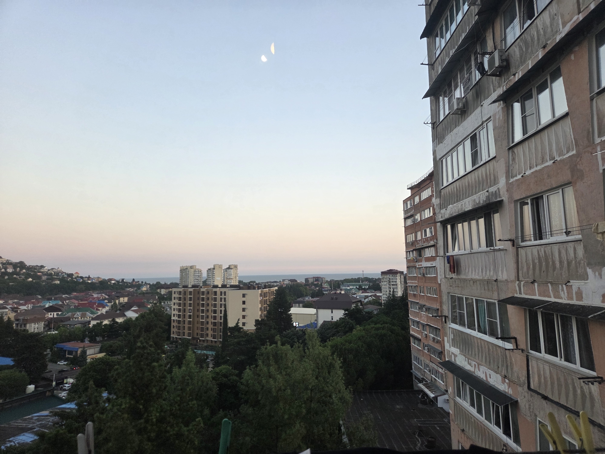 An elevated, wide-angle exterior shot captures a city skyline in Sochi, Russia, at dusk. The foreground is dominated by the right side of a multi-story apartment building with a worn, pinkish-tan facade, featuring numerous windows and balconies. Clothes hang from some balconies. To the left, the scene opens to a densely populated urban area with various buildings, including apartment blocks and smaller houses with colorful roofs, nestled on a hillside. Lush green trees are interspersed throughout the landscape, leading to the background where the Black Sea is visible on the horizon. The sky is a pale gradient of light blue, transitioning to a soft pinkish hue near the horizon, with two crescent moons visible in the upper portion of the sky. The overall impression is that of an urban residential area in the late evening.