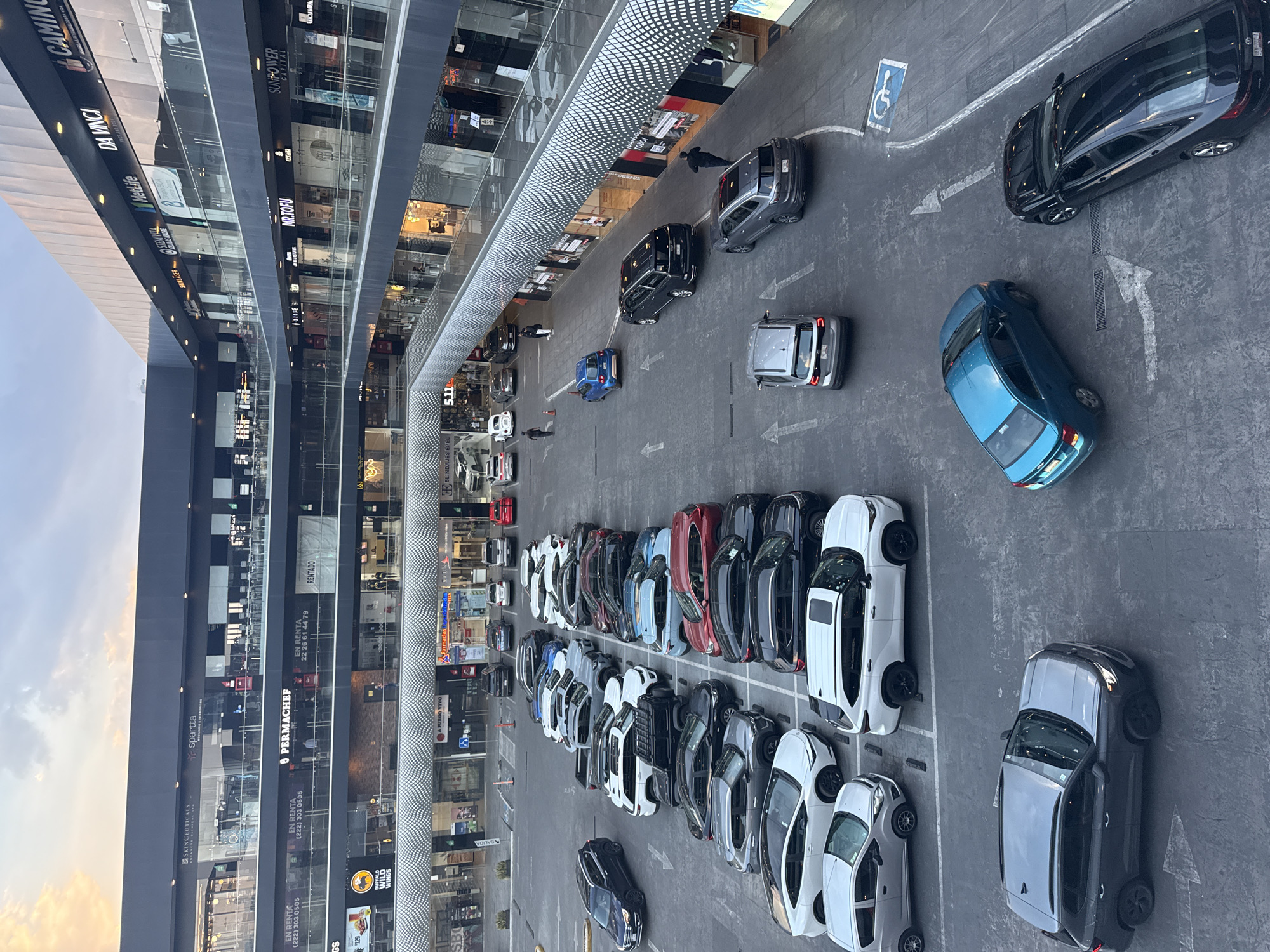 An aerial, tilted photograph presents a vibrant outdoor shopping center in San Bernardino Tlaxcalancingo, Mexico. The scene is dominated by modern, multi-story buildings featuring large, reflective glass facades that house various commercial establishments. A wide paved area in the foreground and mid-ground serves as a parking lot and driveway, filled with numerous vehicles of assorted models and colors, including white, black, grey, blue, and red. Many cars are neatly parked in designated spaces, while others are seen on the road, guided by white directional arrows painted on the asphalt. A blue disabled parking symbol is also visible on the pavement.
The sky in the background appears soft and diffused, indicating either dusk or dawn, though the illuminated storefronts suggest the former. Several individuals can be observed walking near the shops and through the parking area, indicating routine commercial activity.
Prominently displayed text on the building exteriors identifies various businesses and services, including "SKINCEUTICALS," "spartta," "DA VINCI," "MetLife," "MR.TOFU," "PERMACHEF," "SUNPOWER CAPITAL," "5.11 TACTICAL," and "WILD WINGS." Other signs include "EN RENTA" (For Rent) with associated phone numbers (e.g., (222) 303 0505, 22 26 61 44 79), "RENTADO" (Rented), "SALIDA" (Exit), "CLALAS MADRE," "TODO!", and "YPA." The overall impression is that of a bustling, well-maintained commercial complex.