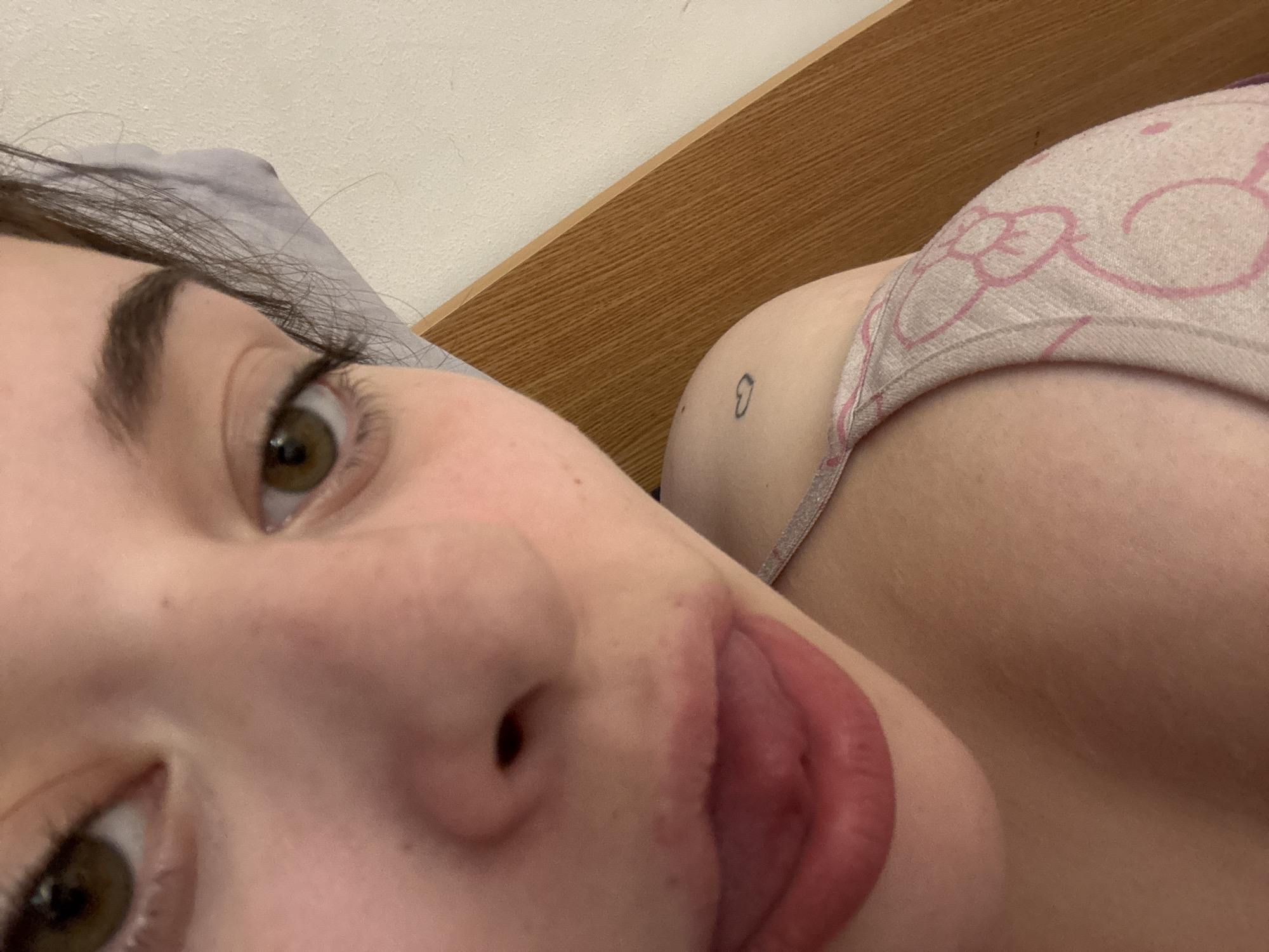 An angled, close-up indoor photograph depicts a person lying down, believed to be in Troyan, Bulgaria. The subject's face is prominently featured in the foreground, with both hazel eyes visible, accompanied by dark eyebrows and eyelashes. The nose and mouth are central, with the tongue slightly extended between red-tinted lips. Dark hair is visible resting against a gray pillow or bedding in the upper left. The upper right portion of the frame shows the subject's shoulder and upper torso, partially covered by a light-colored garment, resembling a bra or bralette, adorned with a repeating pattern of pink bows. A small, black, minimalist tattoo, resembling a tilted heart, is visible on the skin of the upper back/shoulder area. The background includes a light-colored, textured wall and a light-brown wooden headboard with horizontal grain.