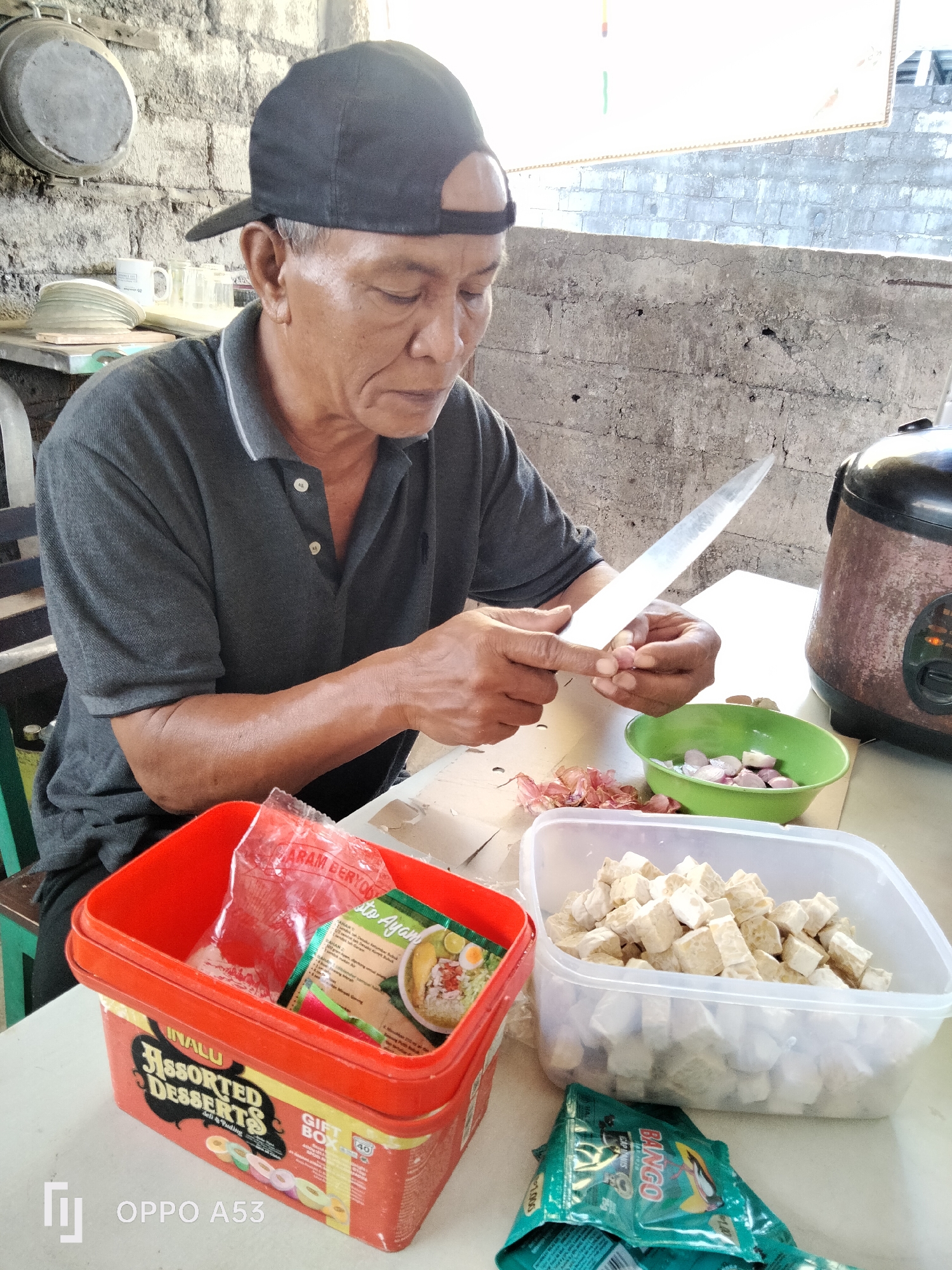 A man wearing a black baseball cap and a dark grey polo shirt is chopping shallots with a knife. He is standing at a table, with various food items laid out in containers. To his right, a green bowl contains more chopped shallots, and a rice cooker is visible in the background. To his left, a red container holds a packet of instant food, likely a soup mix. In front of him, a large plastic container is filled with diced tempeh. The scene appears to be in an outdoor or semi-outdoor kitchen area, with a rough brick wall in the background. The overall impression is of food preparation. The location in Denpasar, Indonesia is suggested by the text "OPPO A53" which is often seen as a watermark on images taken with that specific phone model, and the presence of local food items like tempeh and shallots, commonly used in Indonesian cuisine.