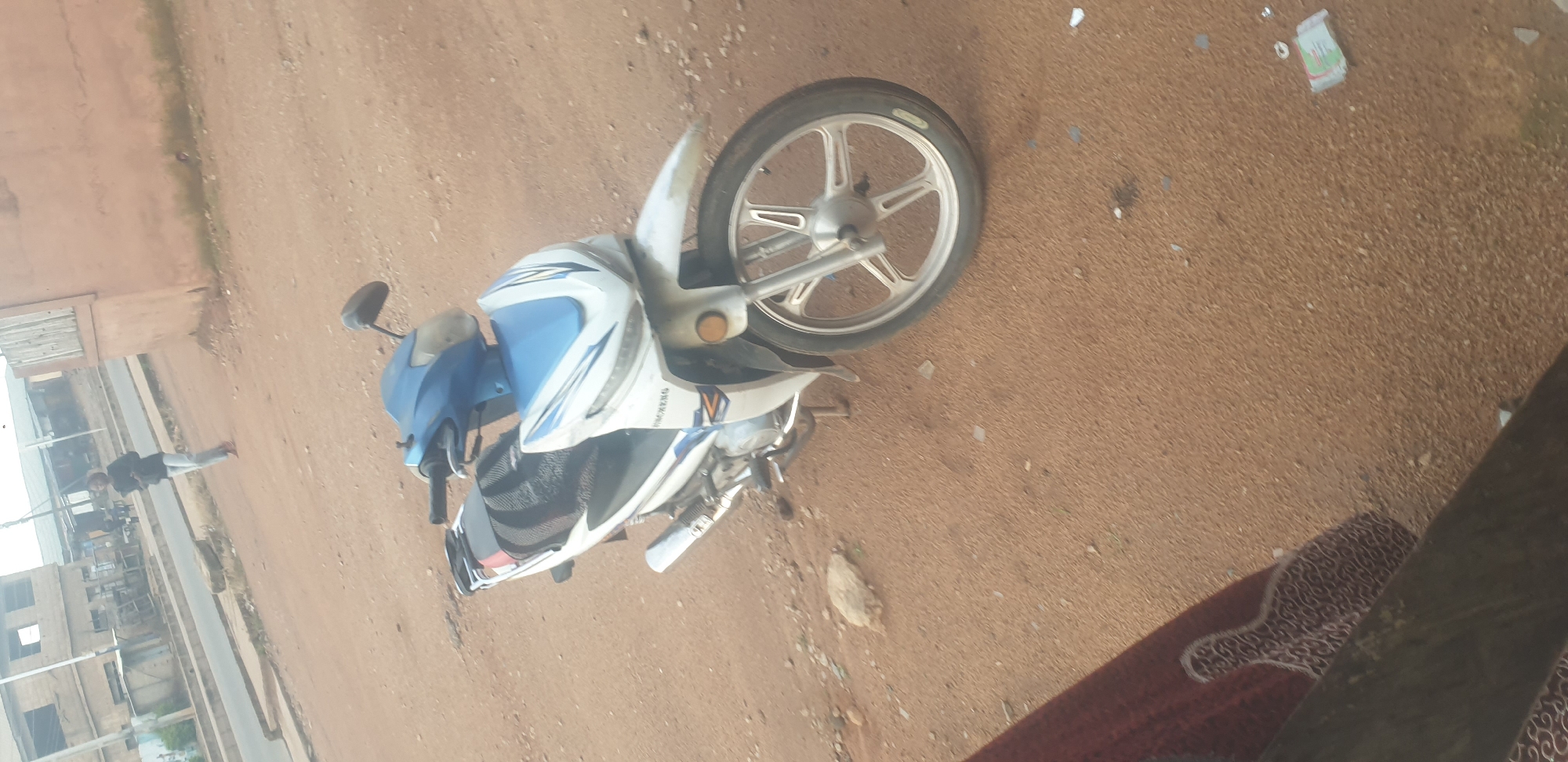 In Jos, Nigeria, a white and blue motorcycle is parked on a dirt surface. The motorcycle has a visible exhaust pipe and a silver rim on the front wheel. In the background, a person is walking on a paved road adjacent to buildings. The environment appears to be an outdoor urban setting with scattered debris on the ground.