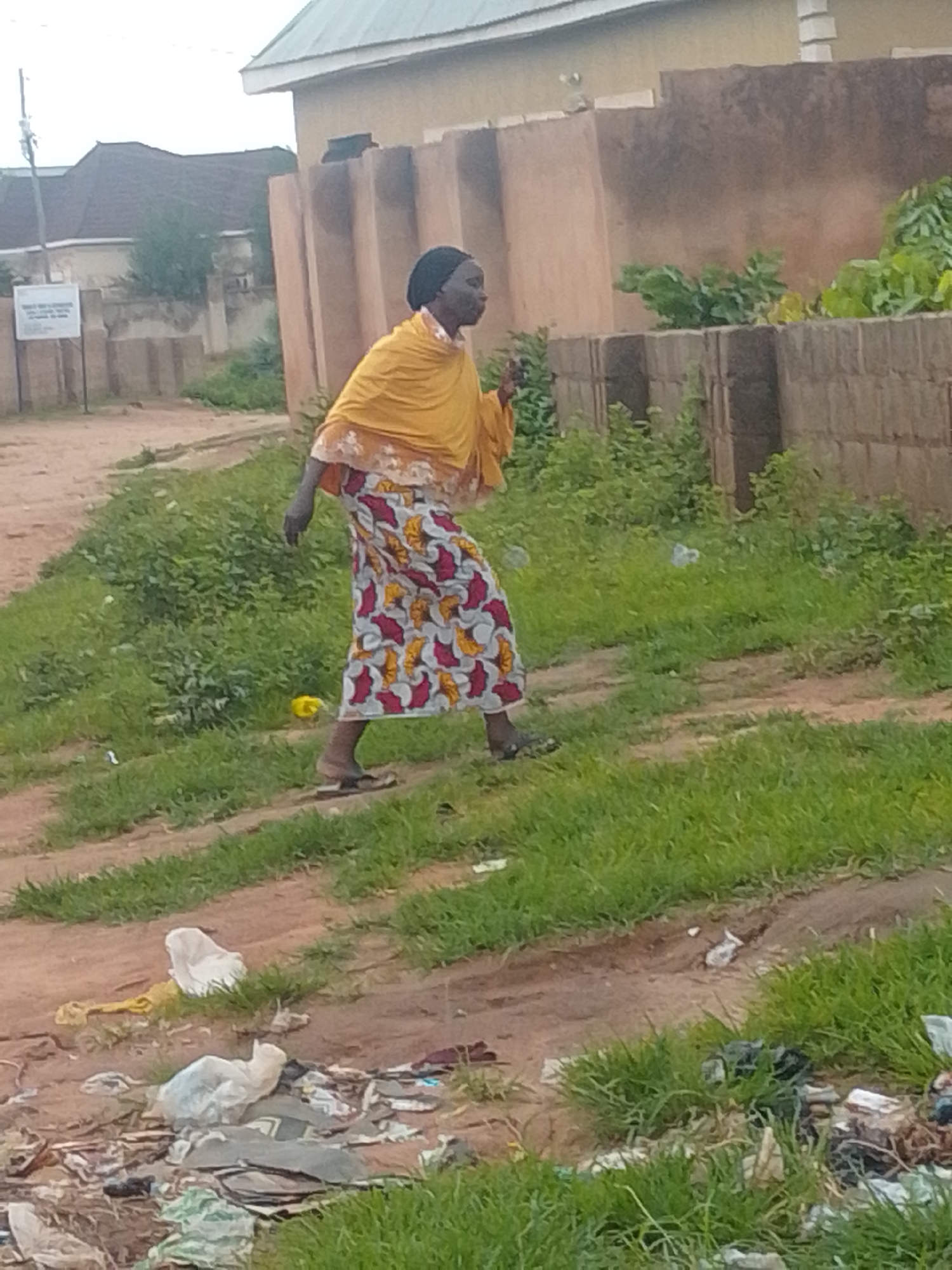 A medium shot shows a woman in profile, walking on a dirt path next to a patch of green grass. She is wearing a yellow headscarf and a patterned skirt with red, yellow, and white designs. Her feet are bare, and she appears to be moving to the right of the frame. Behind her, a weathered brick wall runs horizontally. Beyond the wall, a building with a light-colored roof is visible. In the foreground, the path is littered with various types of trash, including plastic bags and debris. The overall environment suggests a semi-urban or rural setting in Tempure, Nigeria, with a clear contrast between the natural elements, the man-made structures, and the presence of litter.