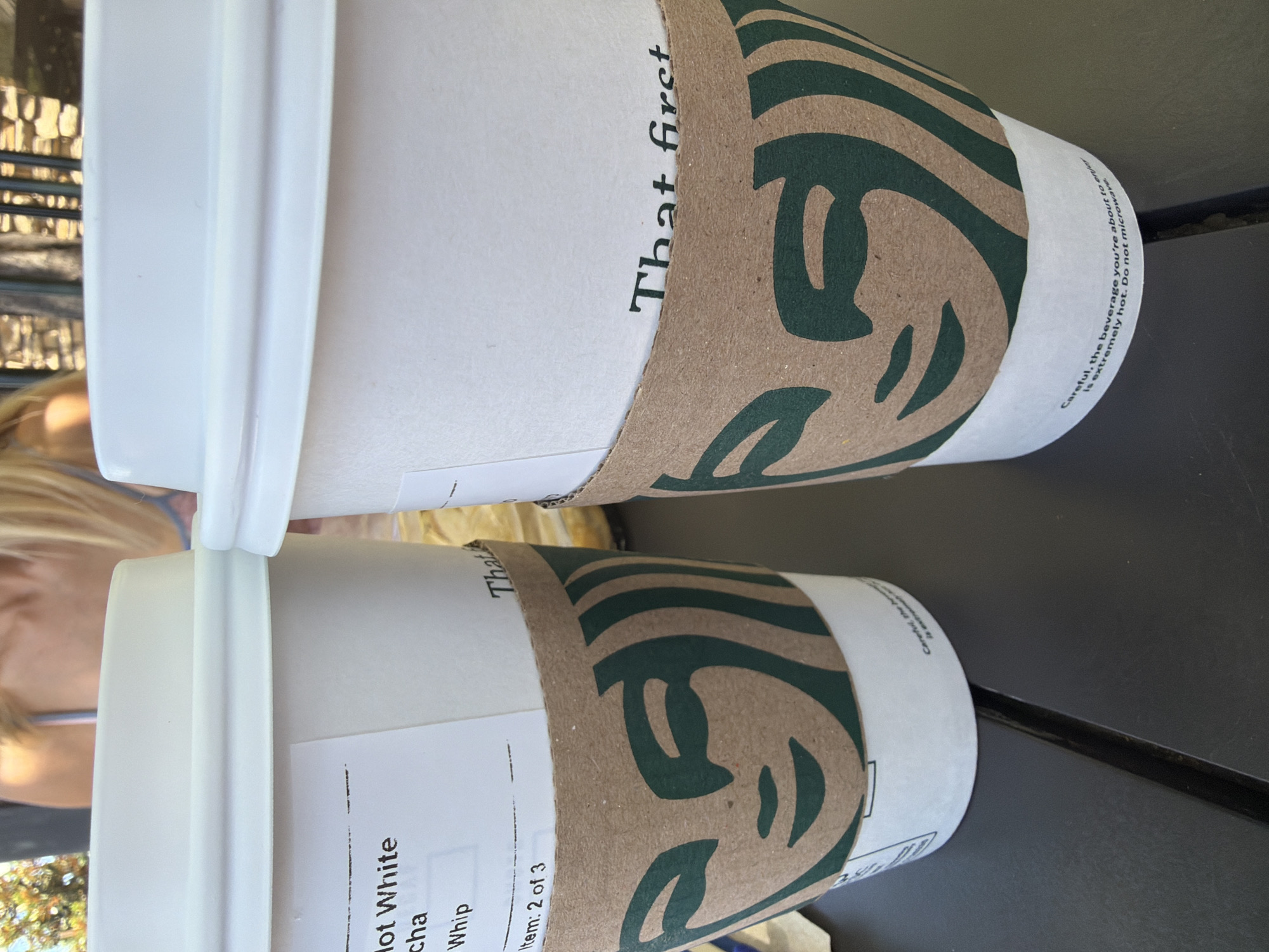 Two Starbucks coffee cups are placed on a dark gray table. The cup on the left has a white label with black text that reads "Hot White Mocha Whip" and "Item: 2 of 3". Both cups have brown cardboard sleeves with the Starbucks siren logo printed in green. The cup on the right has text that reads "That first". The background shows a person with blonde hair and a striped top, and a stone wall behind them. The scene is outdoors.