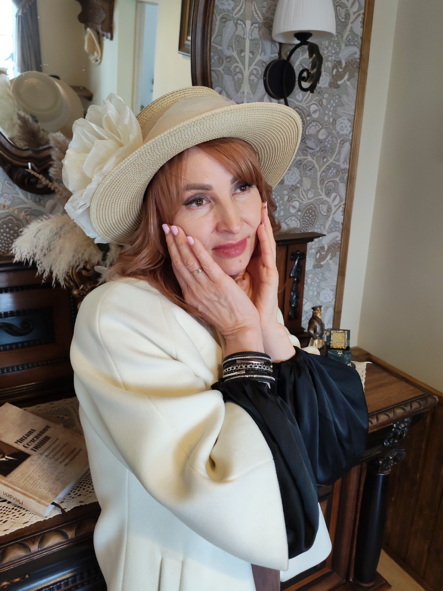 A woman with red hair, wearing a large-brimmed hat with a white flower and a white jacket with black sleeves, is posing indoors with her hands on her face. She is standing near a wooden table with a book on it. Behind her is a mirror reflecting other hats and a wall with patterned wallpaper and a lamp. The image was taken in Sarapul, Russia.