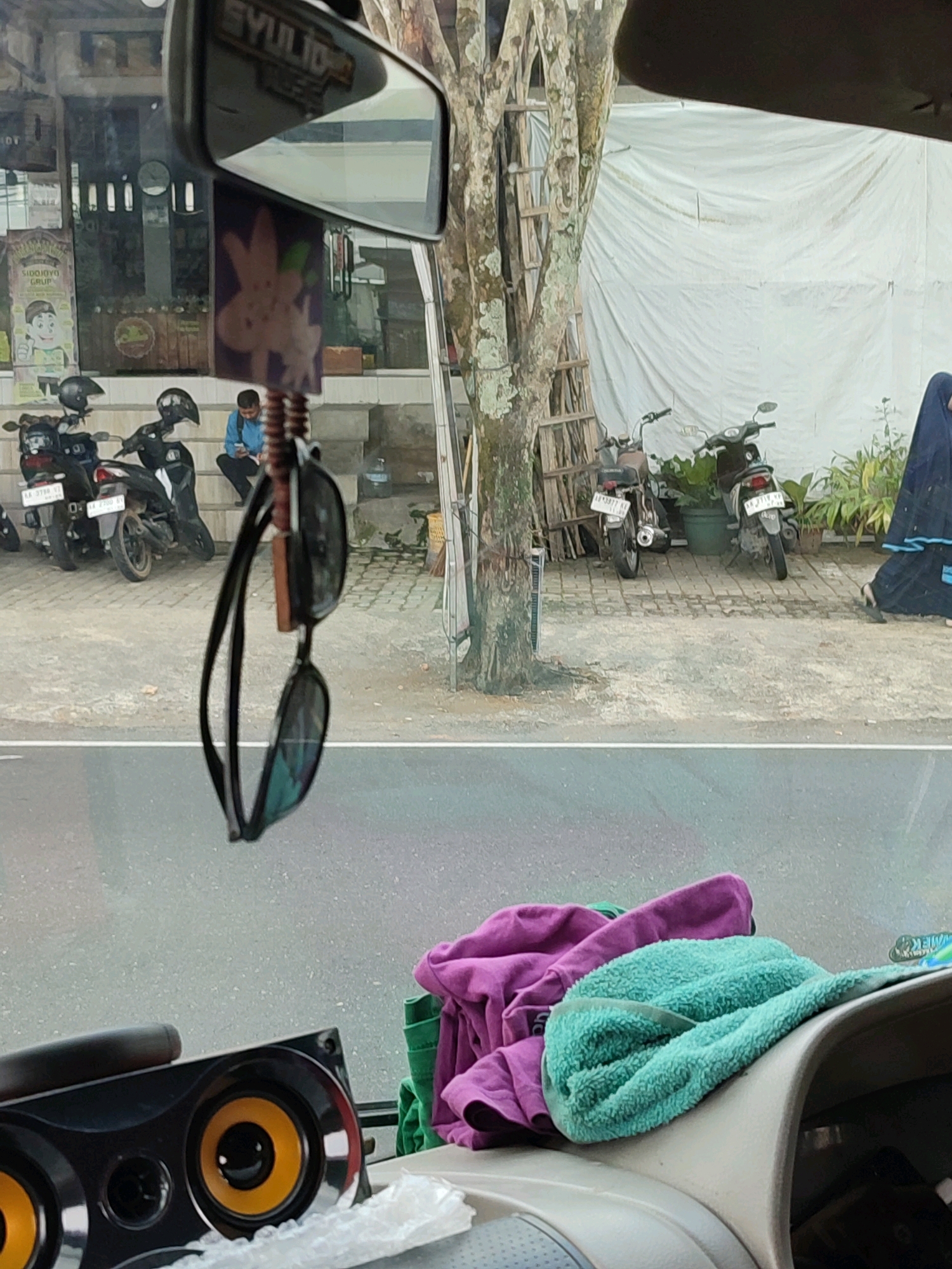 The image displays two scenes, one above the other, suggesting a composite or a view through a vehicle. The upper scene shows a street view in Wonosobo, Indonesia. In the foreground, a pair of sunglasses is hanging from a rearview mirror. Behind the sunglasses, two motorcycles are parked on a tiled surface. A man is seated on a step, looking at his phone. Further back, a white tarpaulin is visible, and to the right, a woman in a dark hijab and dress is walking away. A tree with a ladder leaning against it stands prominently. The lower scene, taken from inside a vehicle, shows a car dashboard with a black speaker on the left. On the dashboard, a pile of clothes, including a purple shirt and a teal towel, is visible. The view outside the vehicle appears to be a road.