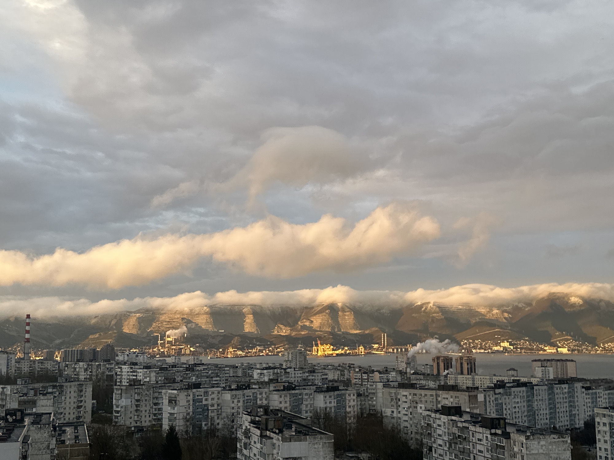 The image presents an elevated view of a cityscape in Novorossiysk, Russia, likely taken from a high-rise building. In the foreground, there are multiple Soviet-era apartment buildings with a repetitive design of rectangular windows and concrete facades. Behind the buildings, a body of water, possibly the Black Sea, separates the city from a range of mountains. The mountains are partially covered in clouds, some of which are illuminated by the sun, creating a dramatic contrast between light and shadow. Several industrial structures, including smokestacks, are visible along the shoreline. The sky is predominantly overcast with a mix of grey and white clouds. No people are visible in the frame. The overall impression is of an industrial port city nestled between the sea and mountains under a cloudy sky.