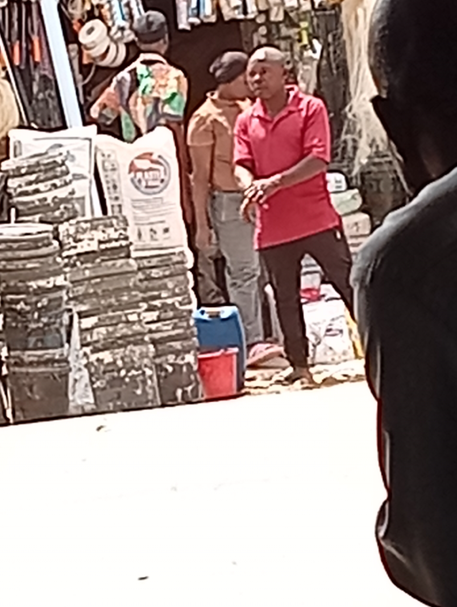 The image depicts a street scene in Maiburiji, Nigeria. There are several people present, including a man in a red polo shirt and black pants standing with his hands clasped, and another individual visible in a brown shirt and jeans behind him. Another person, wearing a colorful shirt, is partially visible. The background includes various objects, such as stacked buckets, sacks labeled "Plastic Steel", and a blue and red container. The environment appears to be a bustling marketplace or street-side shop, with hanging merchandise visible at the top of the frame. The lighting suggests it is daytime. A portion of a person's head and shoulder is visible in the right foreground, partially obscuring the view.