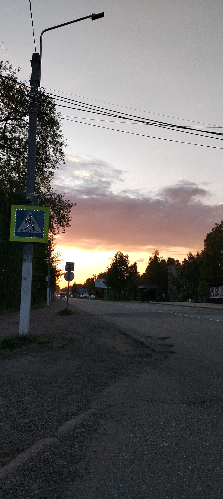 The image shows a street scene in Chaikovsky, Russia at sunset. A pedestrian crossing sign is visible on a metal pole on the left side of the frame, next to a sidewalk with gravel. A road stretches into the distance, and several cars are visible. There is a bus stop and a small store along the side of the road. Trees line the horizon, silhouetted against the orange and purple sky. Power lines stretch across the top of the frame.