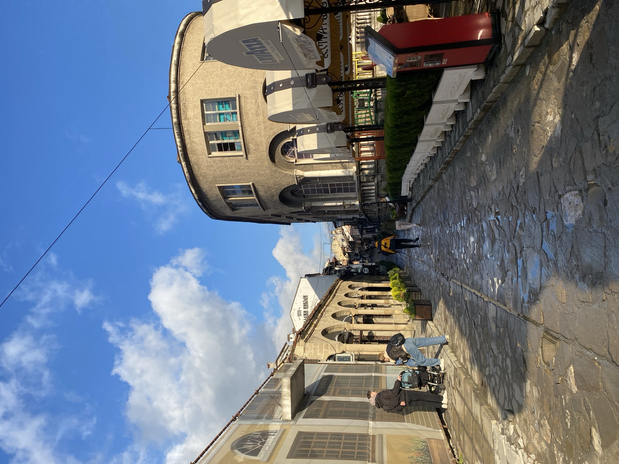 The image depicts a street scene in an unknown city, viewed from an unusual angle. The street, constructed of cobblestones, appears wet. Several people are present, including a group near the bottom of the frame consisting of three individuals and one person walking away further up the street in the distance. Buildings line the street, with a distinctive round structure on the upper end of the frame. The sky is visible, featuring a mix of blue and white clouds. Elements of the environment include awnings, greenery, and other street furniture.