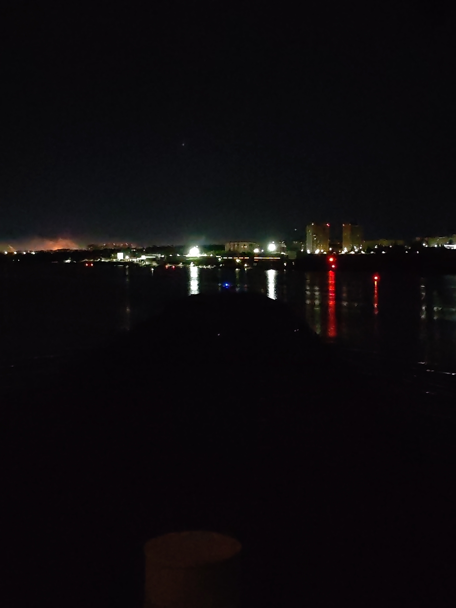 The image depicts a dark, nighttime scene of a city across a body of water. The city skyline is punctuated by bright lights and a few prominent buildings, including two tall buildings with red lights. The water reflects the lights of the city, creating streaks of light in the darkness. The foreground is very dark, obscuring any details. A small light can be seen at the very bottom of the image.