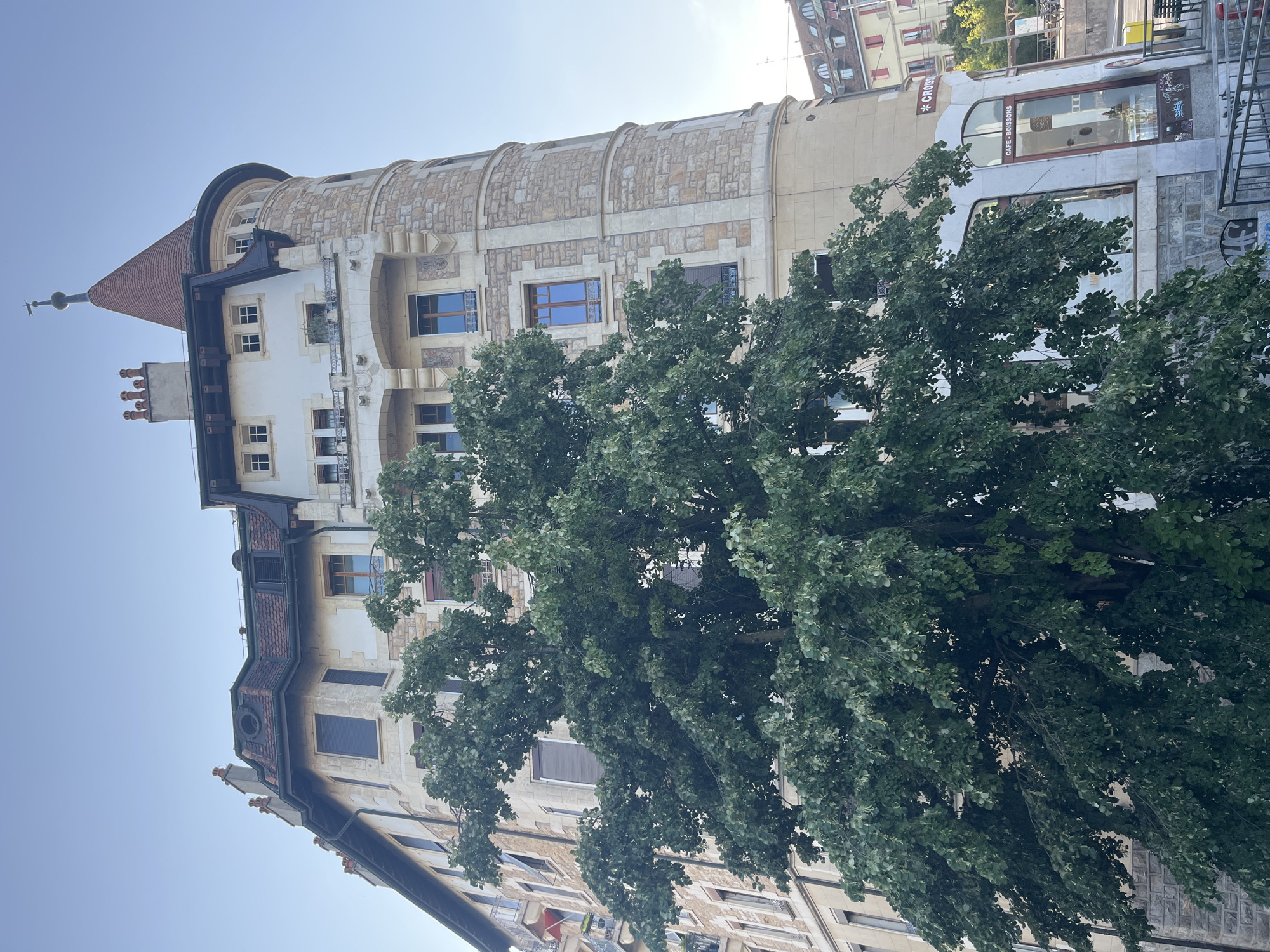 The image shows a building with a turret in Geneva, Switzerland, on a clear day. The building appears to be a residential building with a cafe on the ground floor. A large tree with lush green leaves is in the foreground, partially obscuring the building. The architecture of the building is old European style, with stone and stucco facades, and a red tile roof. The cafe has large glass windows, and signage that reads "CAFE-BOISSONS". The street in front of the cafe has a railing and a yellow bin. In the background, other buildings and trees are visible. The sky is clear and blue.
