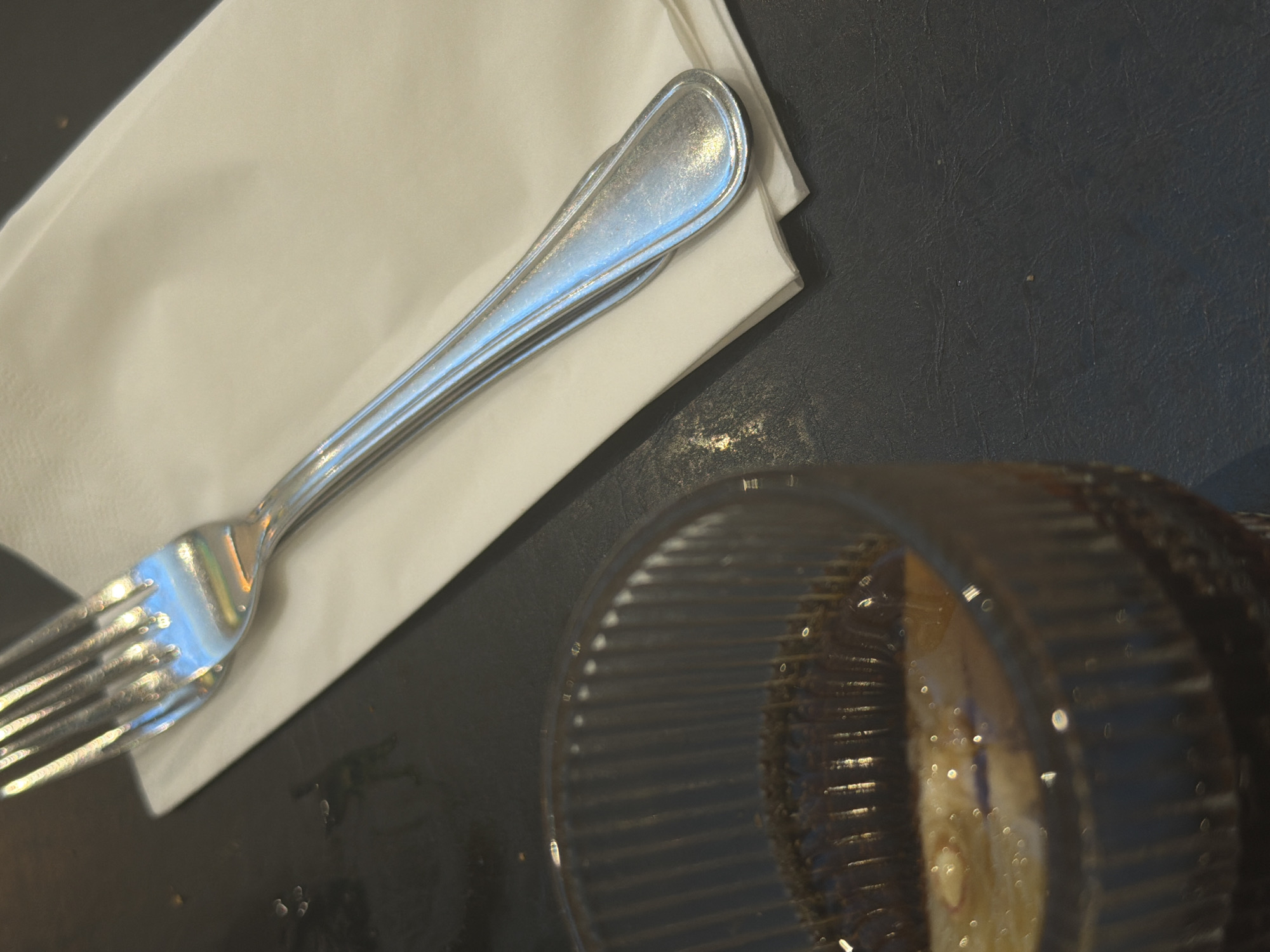 An overhead shot shows a fork resting on a white napkin on a dark, textured surface. To the left, a glass filled with a dark beverage and ice is partially visible. The lighting is bright, creating reflections on the silverware and the glass. The scene is devoid of any human presence.