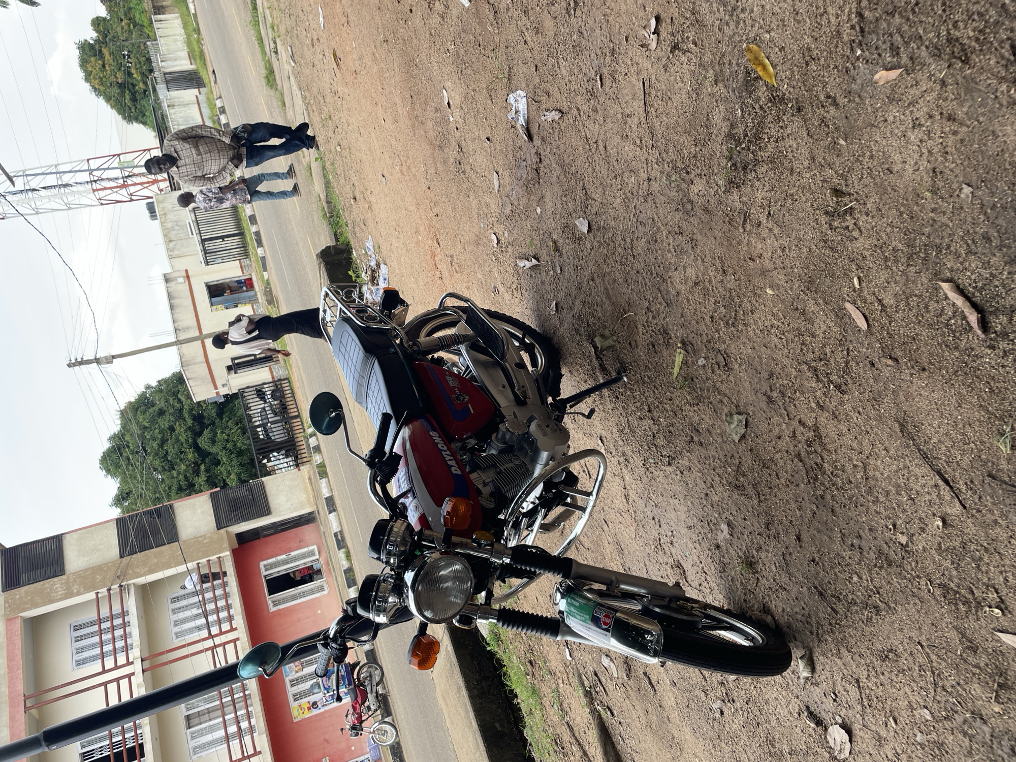 In Jos, Nigeria, a red and white DAYLONG motorcycle is parked on a dirt road beside a multi-story building. The building has several balconies with railings and windows. In the background, several people are walking on the road near a cell tower and a fence. The ground is covered with dirt, leaves, and some litter. The sky is overcast.