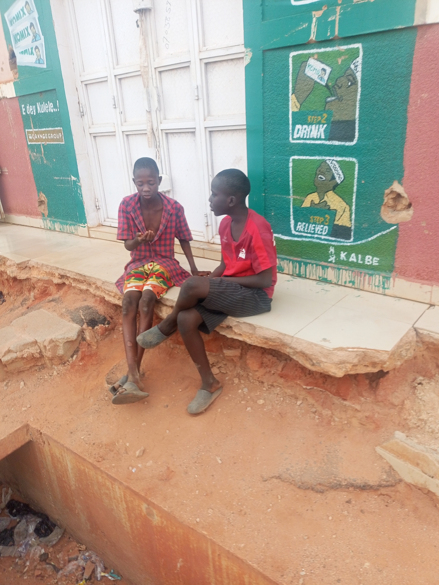 Two young boys are sitting on a raised concrete and tiled platform next to a building wall in Tempure, Nigeria. The boy on the left wears a red and black plaid short-sleeved shirt and multi-colored shorts, while the boy on the right wears a red t-shirt and striped shorts. Both are wearing sandals. They are facing each other, seemingly engaged in conversation or simply resting.
The platform they sit on overlooks a shallow, eroded ditch or drainage channel filled with debris and reddish-brown soil. The background features a light-colored, possibly metal, shuttered doorway or window and a building wall painted in alternating sections of red and teal/green.
The wall is adorned with various texts and illustrations. On the left, "KOMIX" is visible multiple times, accompanied by illustrations of a person. Below this, "E dey Kulele..!" and "ORANGEGROUP" are written. On the right side of the wall, there are two distinct illustrations depicting steps: "STEP 2 DRINK" shows a figure drinking from a packet labeled "Yemola," and "STEP 3 RELIEVED" shows the same figure appearing relieved. Below these illustrations, the word "KALBE" is visible. A faint number "0703857" is also written on the lighter section of the building.
The scene takes place during daylight hours, suggested by the overall brightness, although the exact time of day or specific weather conditions are not clearly discernible. The boys' expressions are neutral.