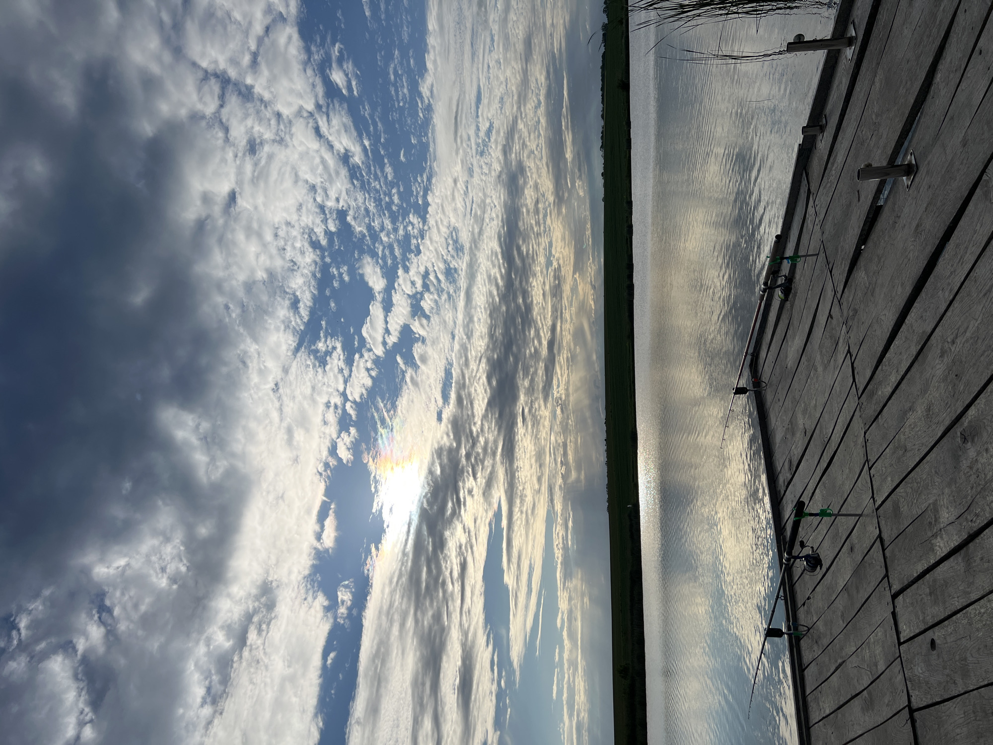 The image depicts a tranquil scene at a lake near Rudky, Ukraine. A wooden pier extends into the water. Fishing rods are placed along the edge of the pier. The lake reflects the sky, which is filled with clouds. A narrow strip of green vegetation is visible on the far side of the lake.