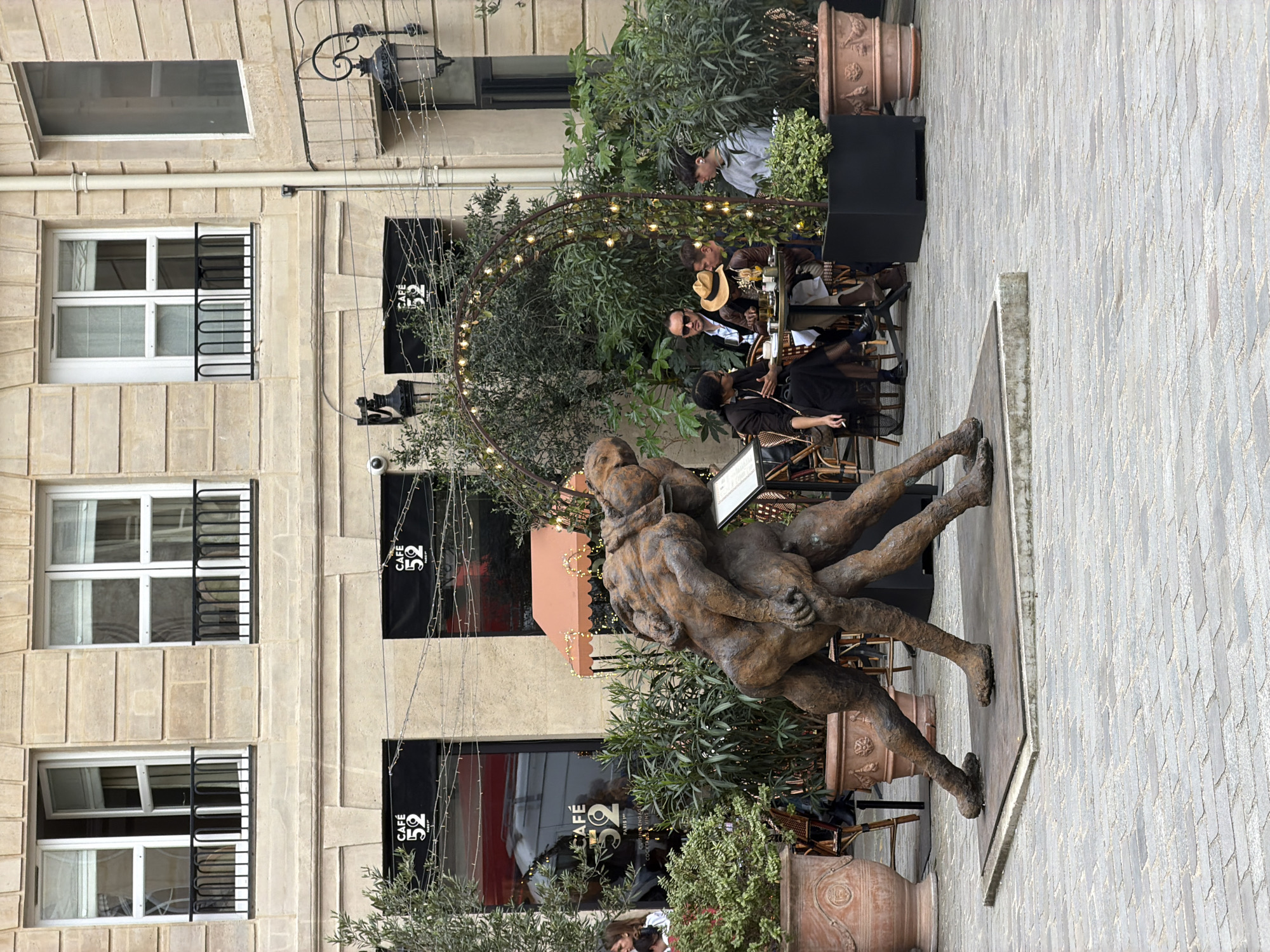 This image captures a vibrant outdoor café scene set on a paved square in Paris, France. A prominent, textured bronze sculpture dominates the foreground, depicting two intertwined human figures in a dynamic pose, possibly embracing or dancing. The sculpture stands on a dark, rectangular base, positioned on light-colored block pavement laid in a herringbone pattern.
Behind the sculpture, an inviting outdoor seating area belongs to "CAFÉ 52," whose name is clearly visible on several black awnings. A diverse group of people are seated at small tables with wicker-style chairs, enjoying drinks and conversation. One person in the foreground right is seen holding a cigarette, contributing to a relaxed, leisurely atmosphere. Others are casually dressed, with one individual sporting a straw hat. The café's outdoor section is enhanced with lush green plants in large terracotta pots and features warm string lights draped over a decorative arched metal structure, suggesting a charming ambiance.
The background is composed of classic multi-story stone buildings, characteristic of Parisian architecture. These buildings feature numerous windows with white frames and decorative black iron railings. An ornate black iron lamp fixture or sign holder is also attached to a building wall. The scene suggests a pleasant day or early evening with mild weather, conducive to outdoor dining and socializing, creating an overall impression of a relaxed and culturally rich urban environment.