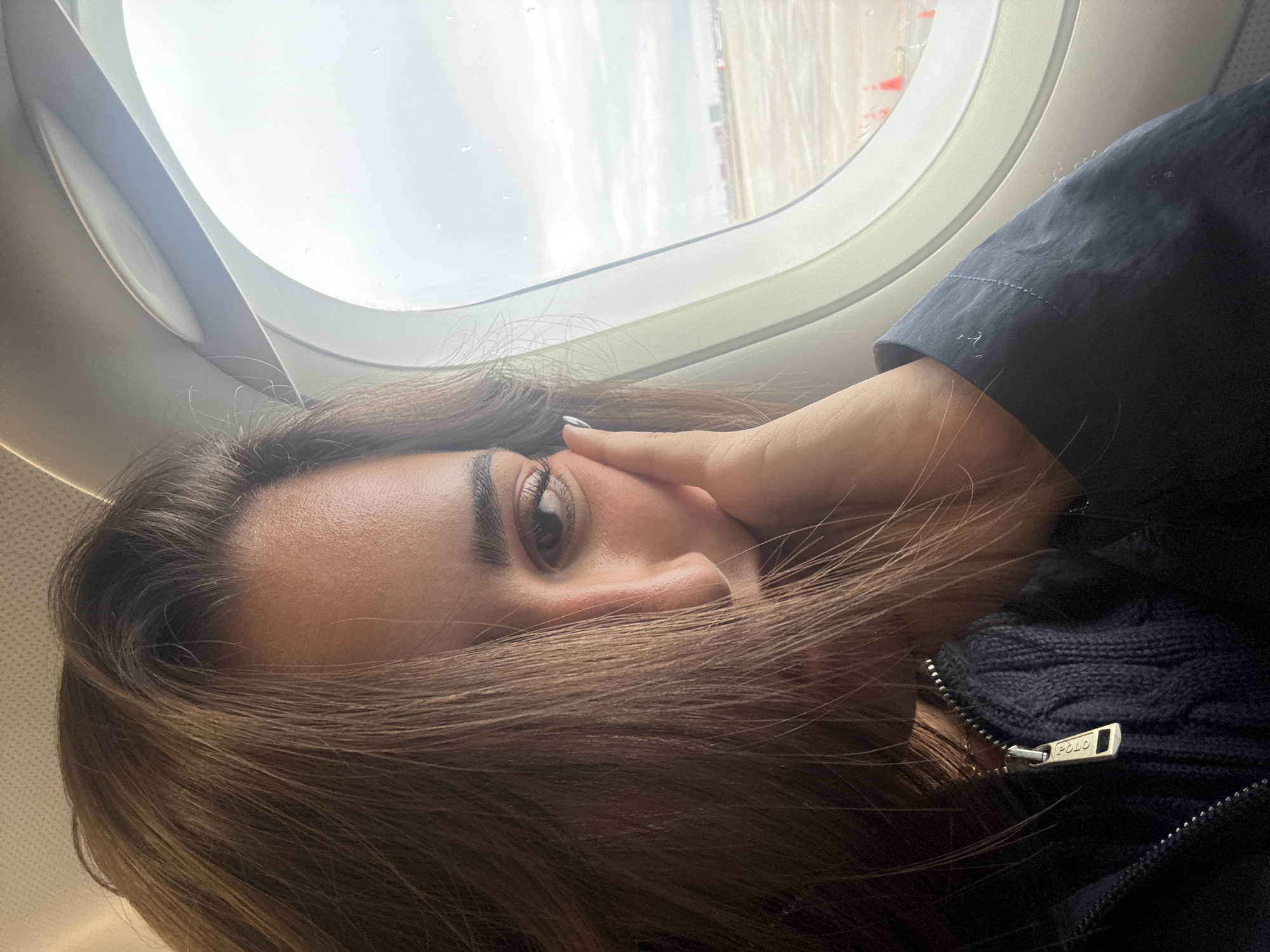 A woman is seated in an airplane by the window. The view outside the window suggests the plane is on the ground or taxiing, with possible airport infrastructure visible. The woman is resting her face on her hand, looking towards the camera. She is wearing a dark jacket or sweater with a visible zipper pull, potentially branded with "POLO". The overall environment appears to be inside an airplane cabin. There is no information to specify its location in San Antonio.