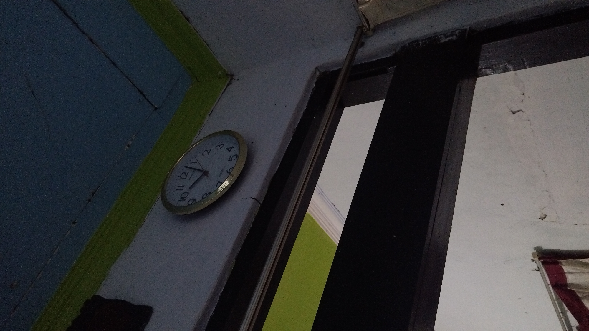 The image shows an analog clock mounted on a light-colored wall. The clock has a golden bezel and black hour and minute hands. The numbers on the clock face are black. The clock is positioned in the upper right quadrant of the image. Above the clock, a blue ceiling with green trim is visible. Below the clock, a dark wooden door frame or window frame is present. The lower portion of the image shows a white wall with some cracking. A portion of a curtain, appearing to be red and beige, is visible in the bottom left corner. The scene is indoors, with artificial lighting creating shadows and a slightly dim atmosphere.