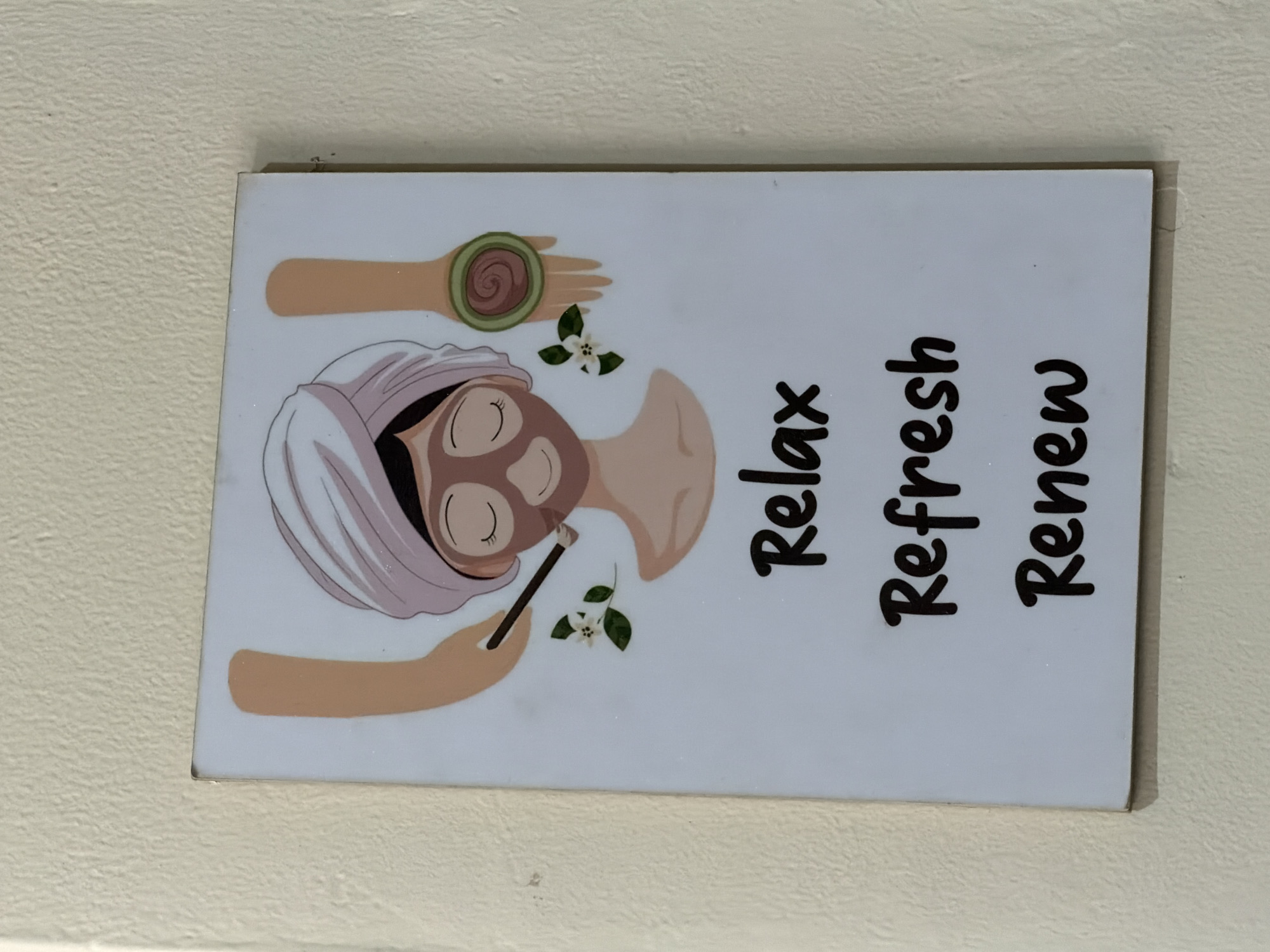 A decorative rectangular plaque, likely made of ceramic or printed acrylic, is displayed vertically against a textured, off-white wall in North Kuta, Indonesia. The plaque features a colorful illustration centered around a spa or self-care theme.
The main subject of the illustration is a cartoon-style depiction of a person receiving a facial treatment. The person has dark hair covered by a light purple towel wrapped around their head, with their eyes closed in a peaceful expression, adorned with long lashes. A brown or clay-colored face mask covers their face. Two light-skinned arms are visible: one holds a brush, applying the face mask, while the other arm extends from the top of the illustration, holding a green and brown swirled object, possibly a rolled-up cucumber slice or a spa product. Small white flowers with green leaves are scattered around the figure, enhancing the natural and calming aesthetic.
To the right of the illustration, three words are written vertically in a black, cursive-like font: "Relax", "Refresh", and "Renew". These words reinforce the serene and rejuvenating atmosphere conveyed by the imagery. The overall impression is one of tranquility and self-pampering. The plaque itself has a slight sheen and a small visible imperfection or chip on its top right edge.