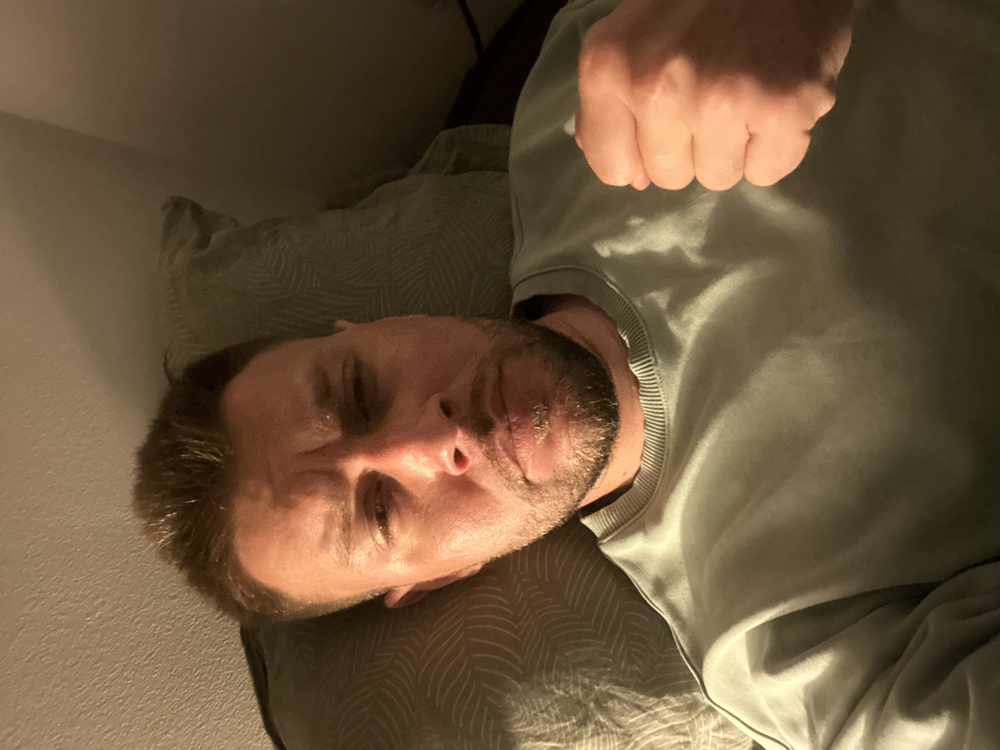 A man is lying on a bed, his head resting on a pillow. He is wearing a light green sweatshirt. His right hand is clenched into a fist and held up near his chest. The lighting is dim, suggesting it may be nighttime or a dimly lit room. The background consists of the pillow and a textured wall. The man's expression is serious, with his brow furrowed. No specific location in Zvezdara Urban Municipality, Serbia, can be identified from the image.