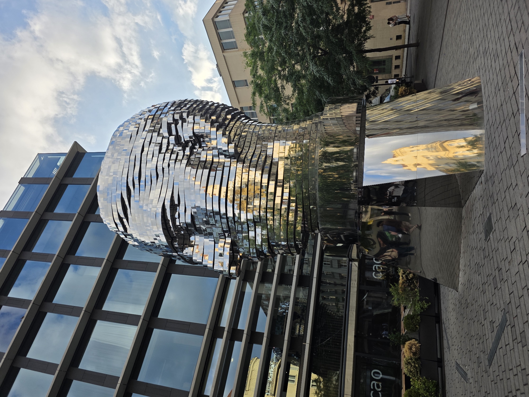 This is an outdoor, eye-level, full shot of a large, reflective sculpture of a human head. The sculpture is composed of many horizontal, wavy metallic strips that create a fragmented, mosaic-like effect. The sculpture is located in front of a modern building with a glass facade. The building's windows reflect the sky, clouds, and the surrounding environment, including the sculpture itself.
To the right of the sculpture, there is a pavement made of gray bricks. In the background, there are more buildings, some with trees and foliage in front of them. People are visible walking on the pavement and reflected in the sculpture and the building's facade. The scene appears to be set during the daytime, with natural light casting reflections. The sky is partly cloudy. There is some visible text on the glass facade of the building, including "cao" and "galeria," which suggests a retail or gallery space. The overall atmosphere is urban and contemporary, highlighted by the striking sculpture and modern architecture.