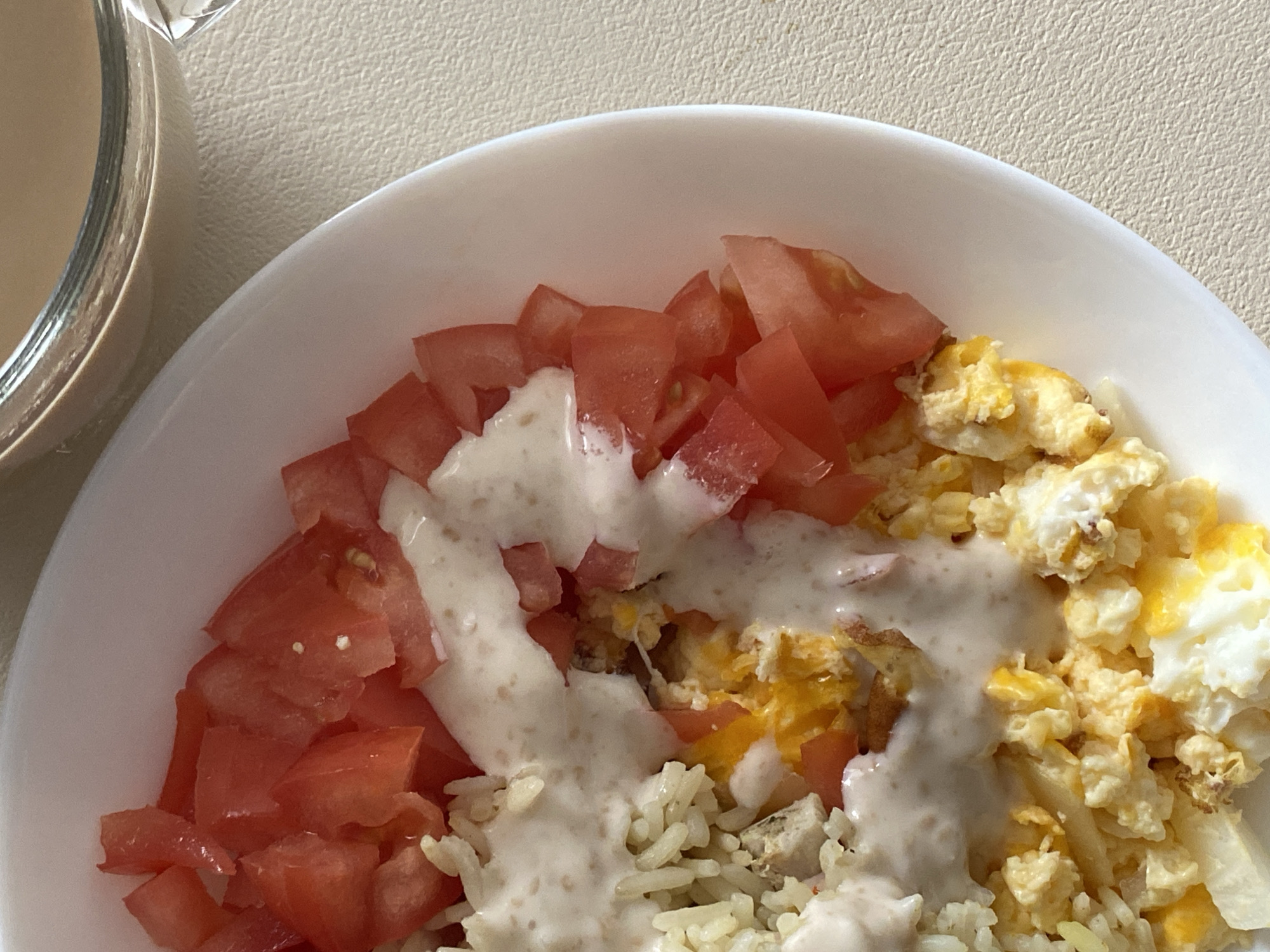 A close-up overhead shot shows a white bowl filled with a meal. The bowl contains chopped tomatoes, scrambled eggs, rice, and a creamy white sauce. To the upper right of the bowl, part of a clear glass containing a light brown beverage is visible. The meal is presented on a textured, light beige surface.