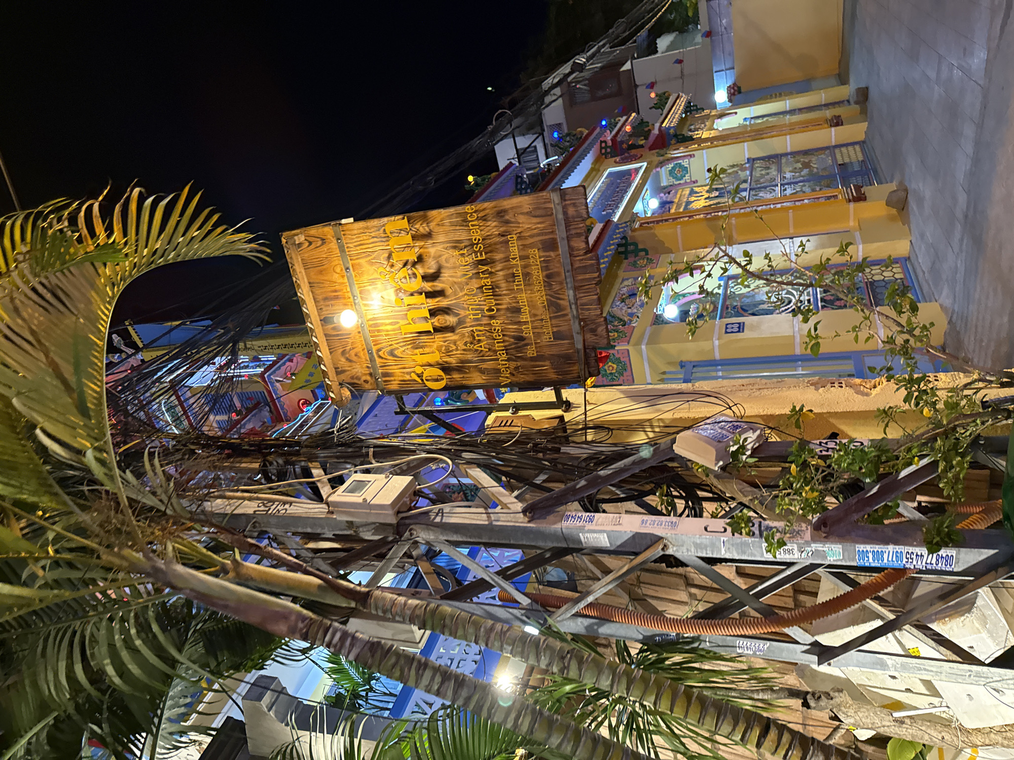 This is a nighttime exterior shot of a restaurant named "Om Hiếm" in Nha Trang, Vietnam. The restaurant's sign, made of wood with the name in yellow letters and a smaller English description "Vietnamese Culinary Essence," is illuminated by a light bulb. Below the sign, the entrance to the restaurant is visible, featuring bright yellow walls, blue gates with intricate designs, and colorful decorative elements. A palm tree is prominently featured in the foreground, with its fronds extending towards the top left of the frame. A complex network of electrical wires is also visible, attached to a utility pole in front of the building, partially obscuring the restaurant's facade. The pavement in front of the restaurant is visible, as is a portion of another building on the right side of the frame.
