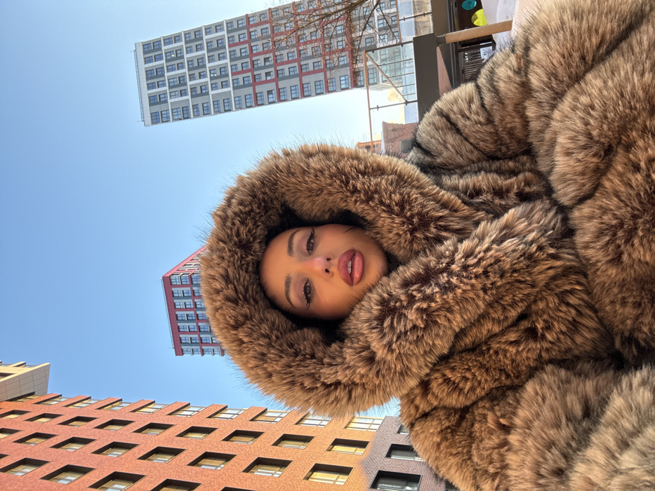 A medium-shot selfie captures a woman outdoors in the ZILART residential complex in Moscow, Russia. The central subject is a woman from the chest up, wearing a voluminous, hooded faux fur coat in shades of brown and tan. Her hood is up, framing her face, which features dark hair, prominent eyeliner, dark eyebrows, and plump lips accentuated with pinkish-red lipstick, slightly parted. She maintains direct eye contact with the camera. The background reveals a clear blue sky and three distinct modern high-rise buildings. To the left, a multi-story building with a reddish-brown brick facade is partially visible. In the middle-rear, a reddish-pink apartment building stands. To the right, a tall, slender residential tower with a grey and white facade featuring red window accents dominates the skyline. Snow covers the ground in the foreground and beneath the buildings. A glass barrier is partially visible behind the woman to the right. The scene is brightly lit by direct sunlight, suggesting a clear, cold-weather day.