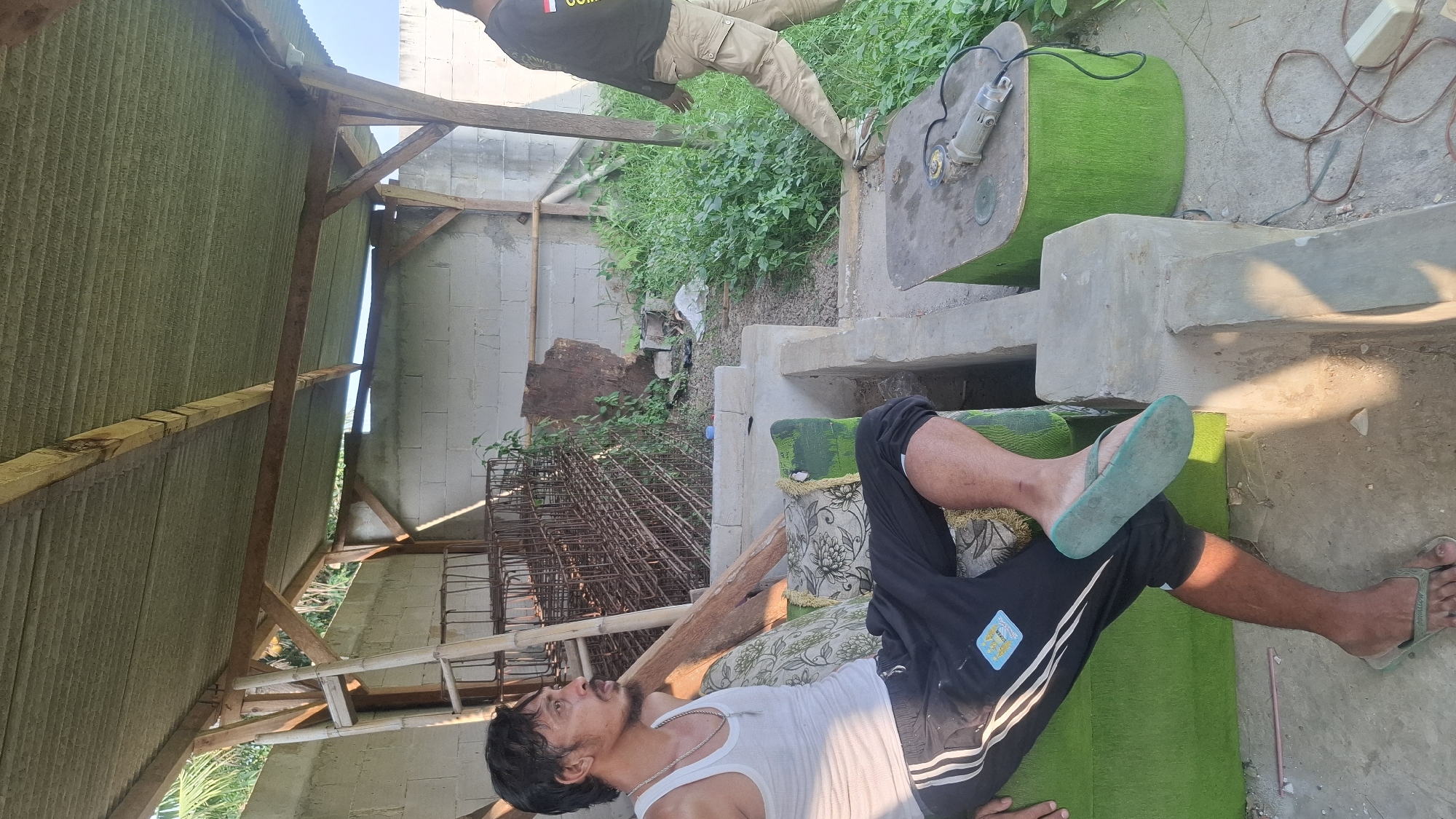 The image depicts an outdoor scene in what appears to be a semi-constructed or utilitarian area. In the foreground, a man with dark hair and a mustache is seated on a green upholstered bench. He is wearing a white tank top, black track pants with white stripes, and teal flip-flops. His left leg is crossed over his right, and his feet are resting on the ground and the bench. Behind him, a stack of metal rebar cages is visible, along with a wooden ladder leaning against a support beam.

To the right of the man, a small green table holds a silver angle grinder with a black power cord. Further to the right, another person is walking away from the camera, wearing a dark long-sleeved shirt and light-colored cargo pants. The background consists of a grey concrete block wall, overgrown green vegetation, and a corrugated metal roof supported by wooden beams, suggesting an unfinished shelter or workshop. The ground is a mixture of concrete and dirt, with some debris scattered around. The overall impression is one of a rural or workshop environment in Rahong, Indonesia, with a focus on manual labor or construction activities.