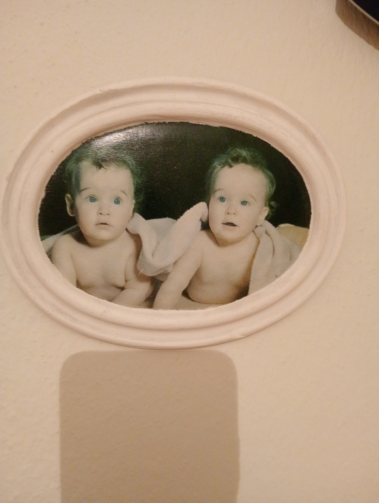 The image is a photograph of a framed oval portrait of two babies, likely twins, lying on their stomachs and wrapped in a light-colored blanket. The babies have light skin and blue eyes, with sparse dark hair. The left baby is looking directly at the camera with a slightly surprised expression, while the right baby is looking slightly to the left. Both babies are shirtless. The portrait is framed by a light-colored, decorative oval mat. The background within the frame is dark. The outer wall surrounding the frame is a light, textured cream color, with a distinct shadow cast to the left of the frame. The photograph appears to be taken indoors. The location of this image in Silifke, Turkey is not discernible from the visual information provided in the photograph.