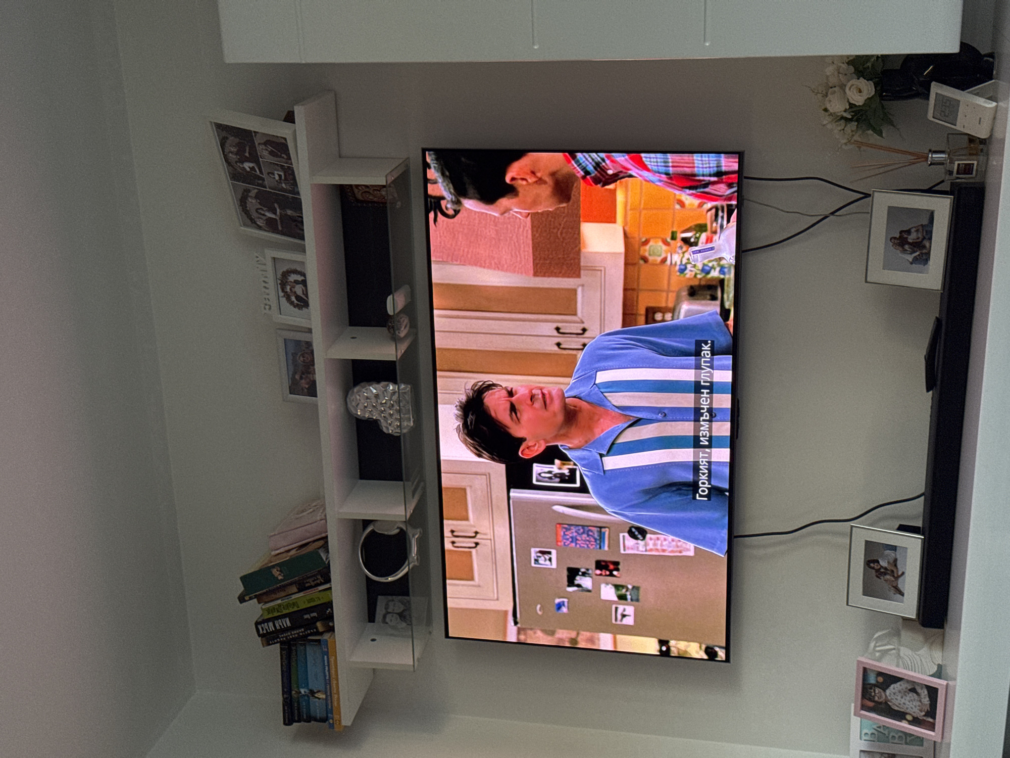 A tilted medium shot depicts a television mounted on a wall, displaying a scene from the sitcom "Two and a Half Men". The characters in the scene are Charlie Harper (played by Charlie Sheen) and Alan Harper (played by Jon Cryer). The language on the screen is Bulgarian. The refrigerator behind the characters is adorned with photographs and magnets. To the left of the TV is a white wall-mounted shelf containing books, decorative items, and framed pictures. To the right of the TV are framed pictures, a flower arrangement, an air freshener, and a sound bar. Given the Bulgarian language on the screen, the scene is likely located in Sofia, Bulgaria.